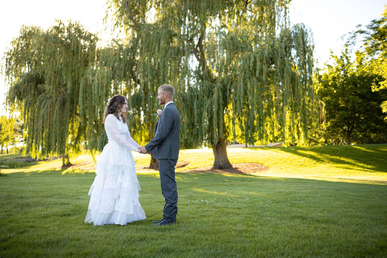 Couple in wedding attire standing together on green lawn with willow trees at sunset.