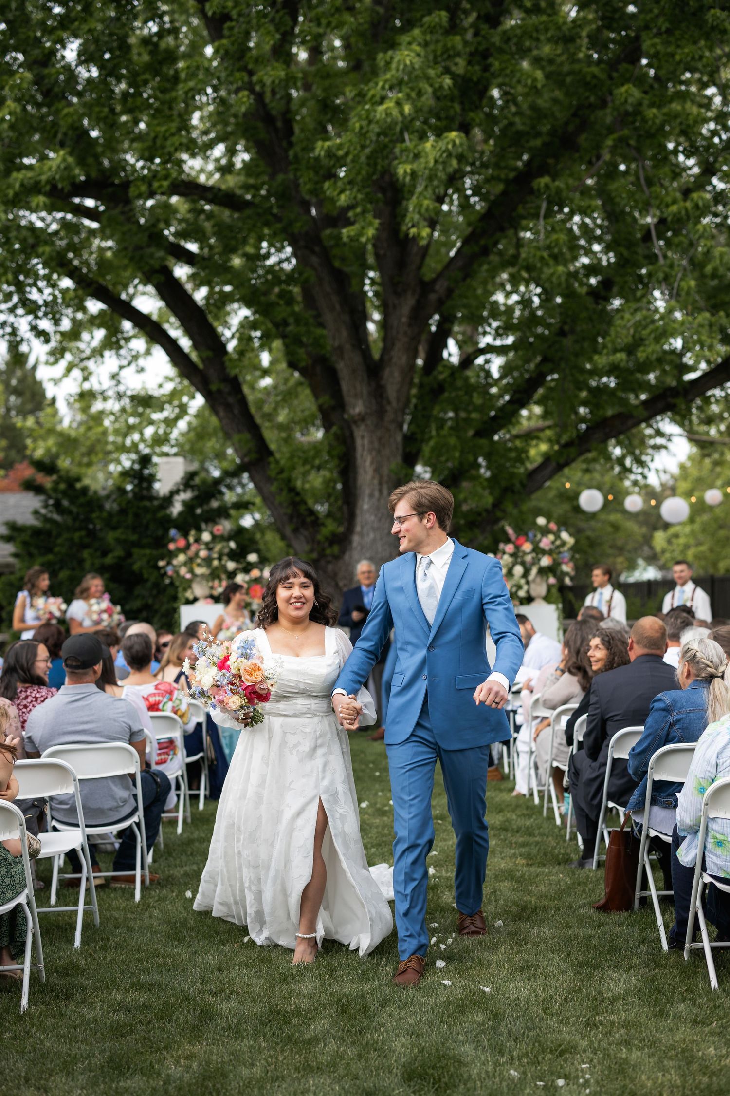 Joyful couple walking down aisle between white chairs at outdoor garden wedding.