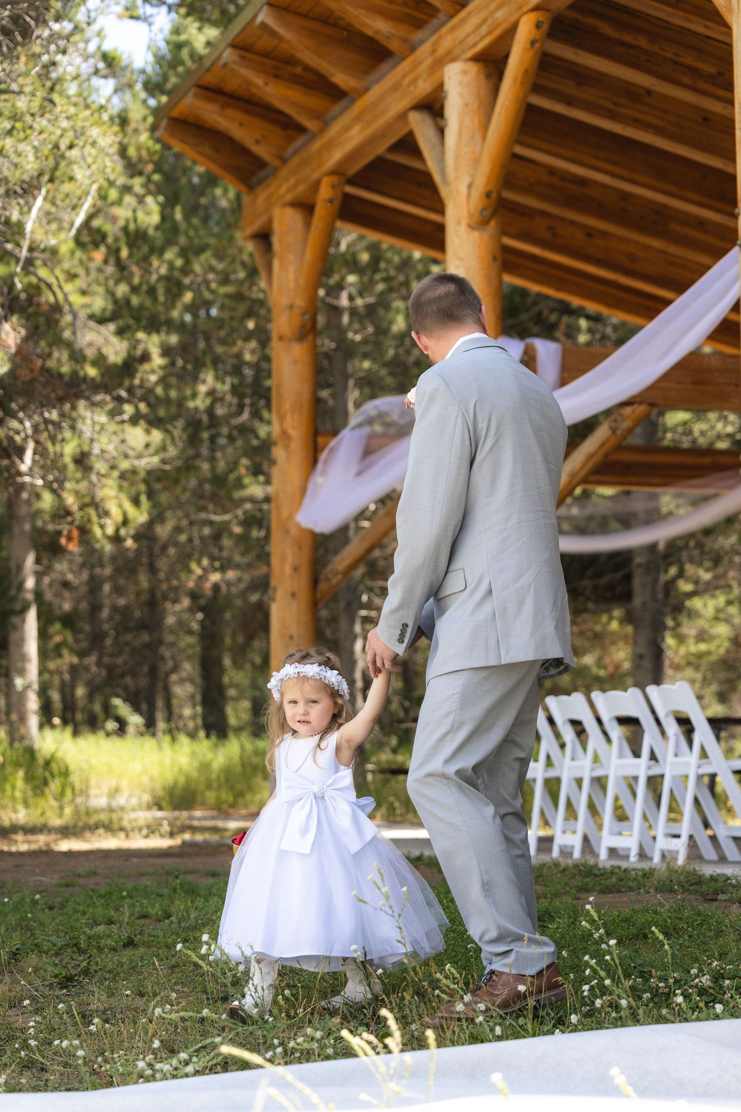 A person in grey suit dancing with small child in white dress under rustic wooden pavilion.