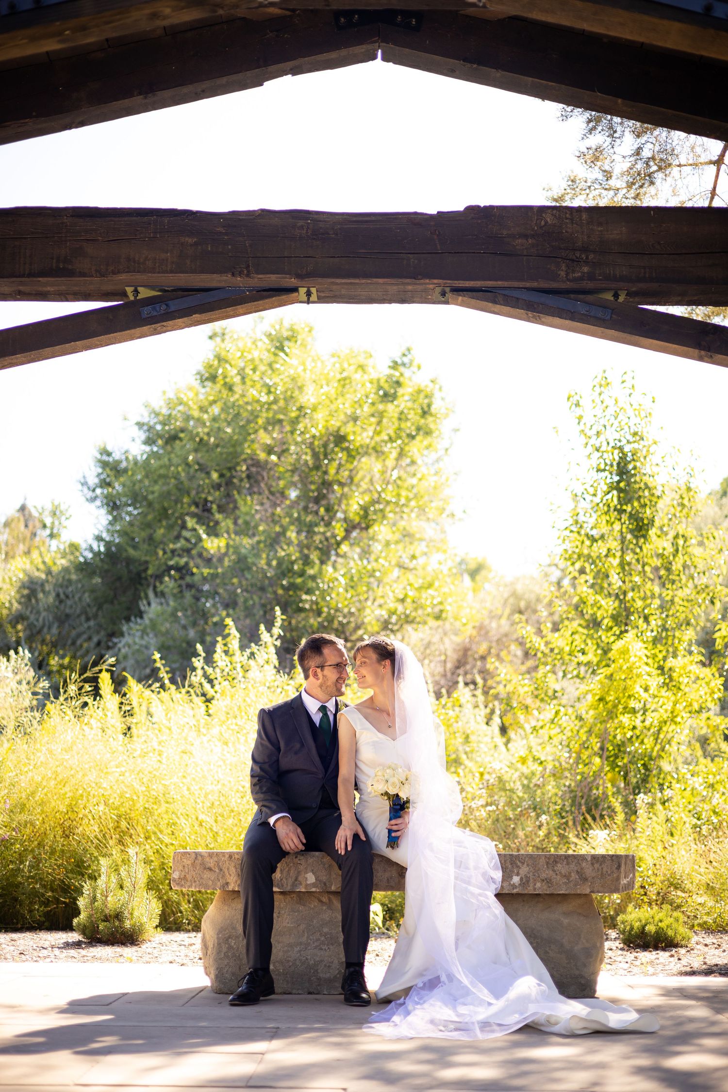 Couple in formal wedding attire sits together on stone bench under wooden beams in outdoor garden setting.