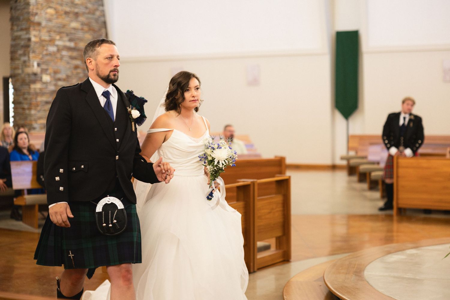 A couple walks down the aisle during their wedding ceremony, with the groom wearing a traditional Scottish kilt.
