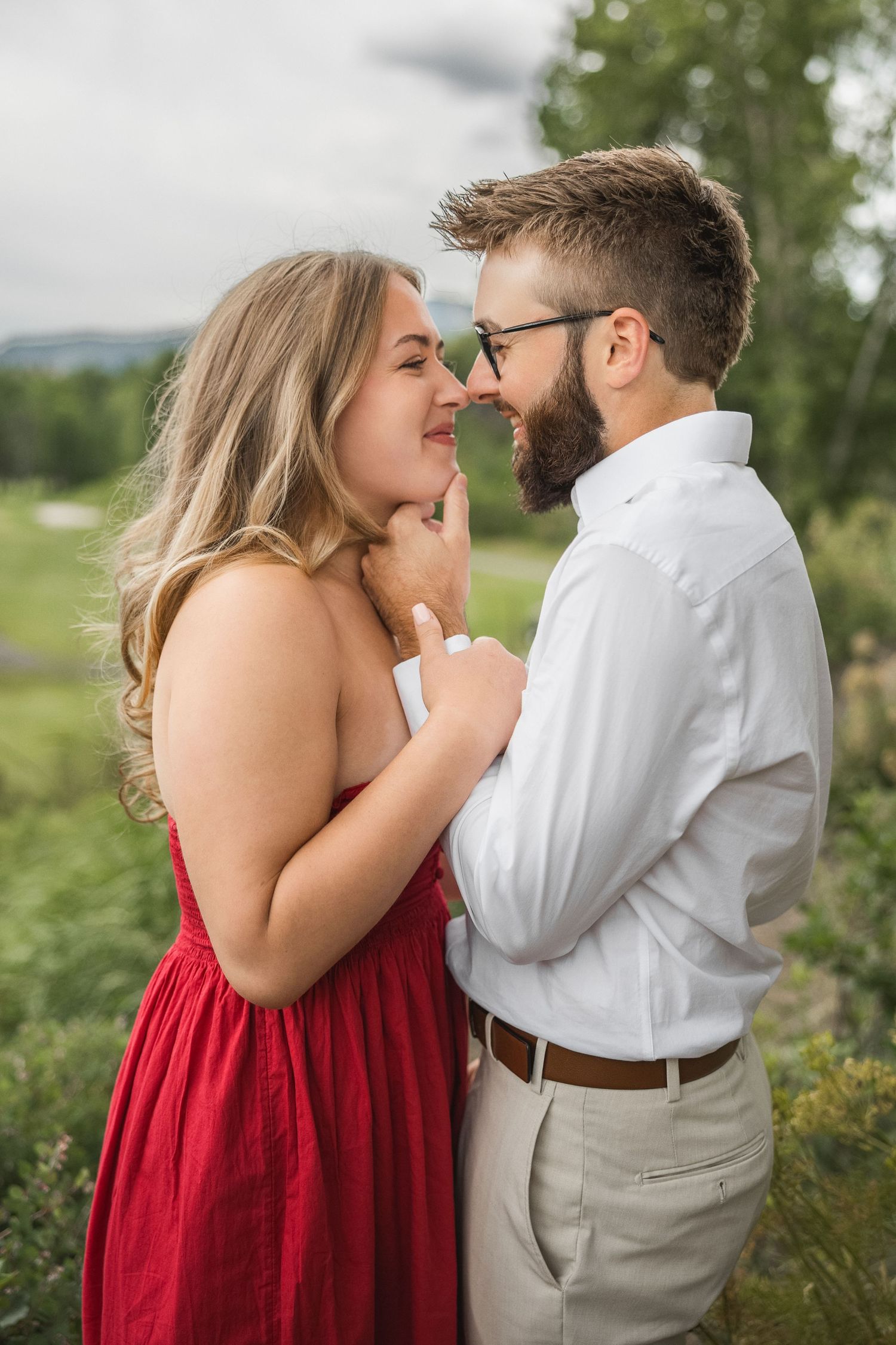 A romantic couple shares an intimate moment outdoors, with her in a red dress and him in a white button-down shirt.
