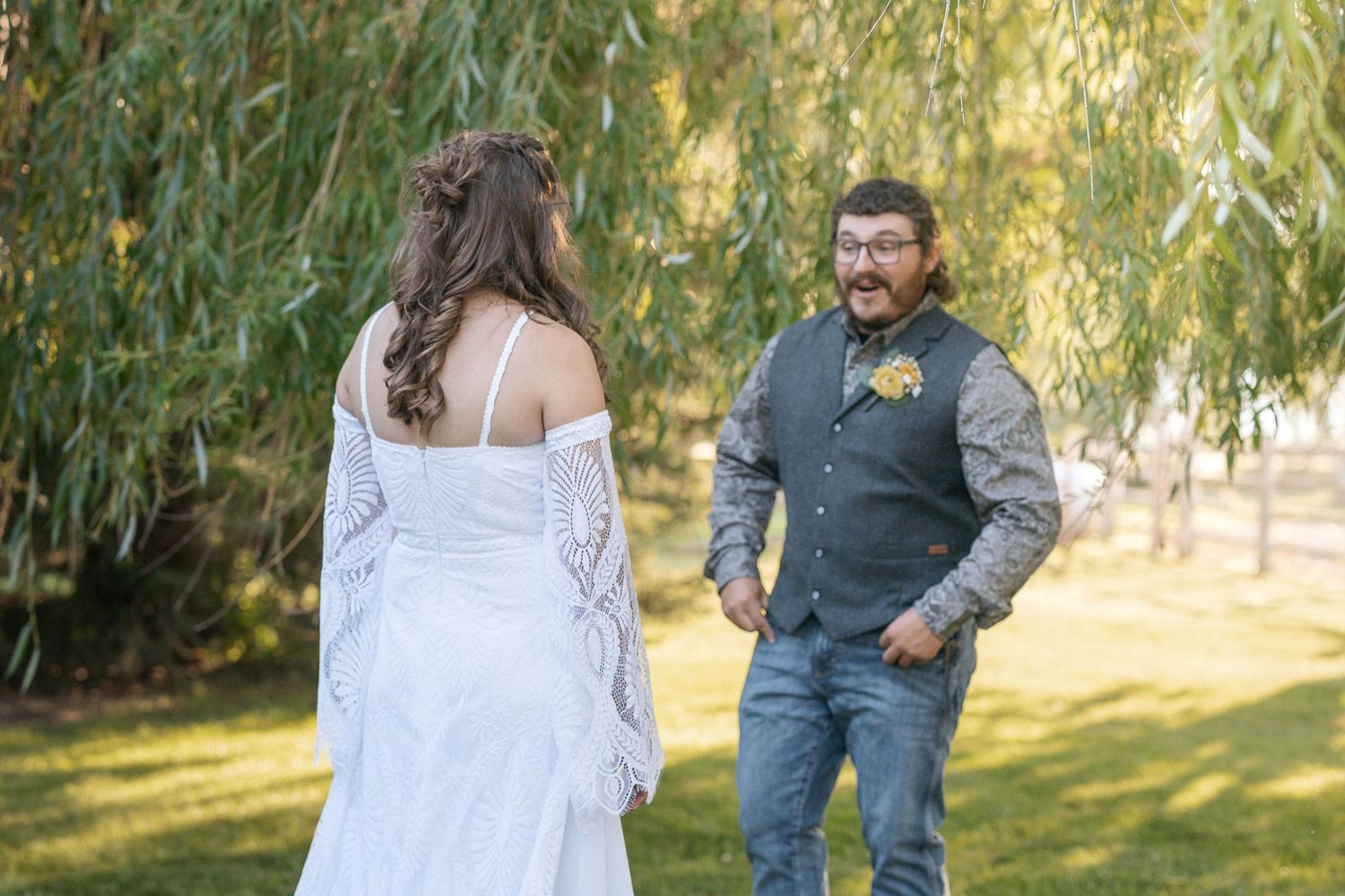 A couple in wedding attire shares a romantic moment under sunlit willow trees in a garden setting.