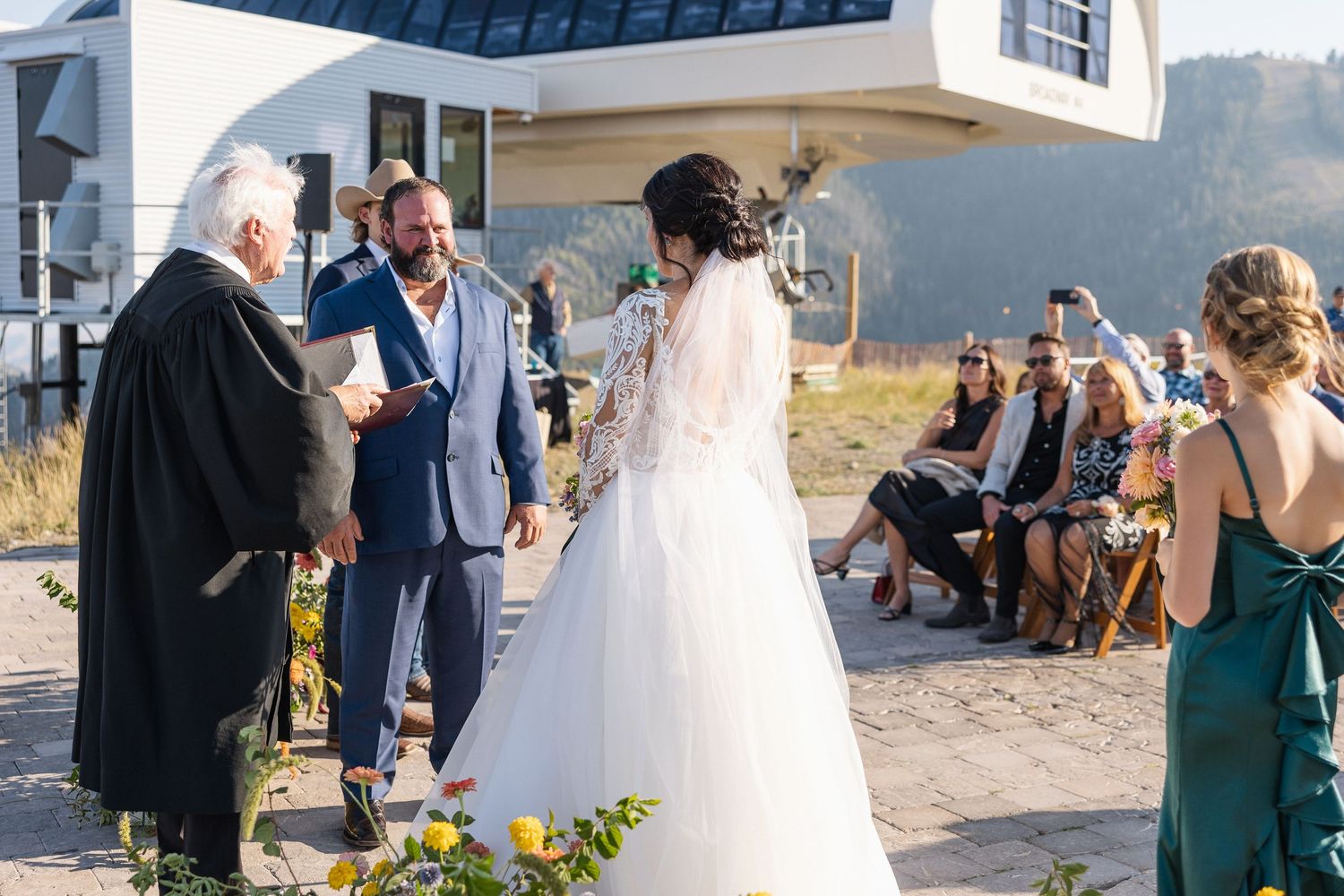 A wedding ceremony takes place outdoors with guests seated and an officiant leading the service in front of mountain views.