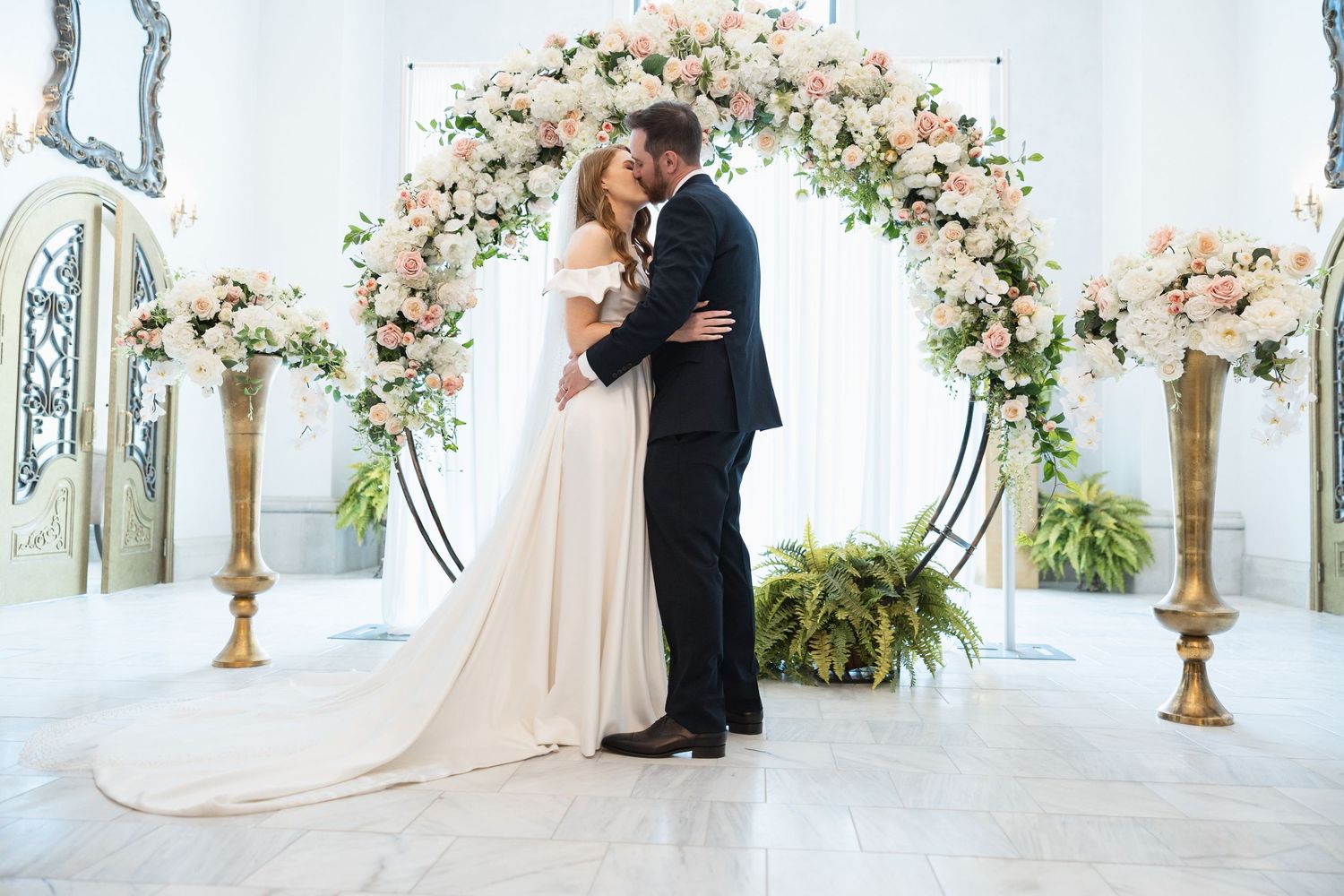 Wedding ceremony in progress under ornate floral arch with blush and white flowers.