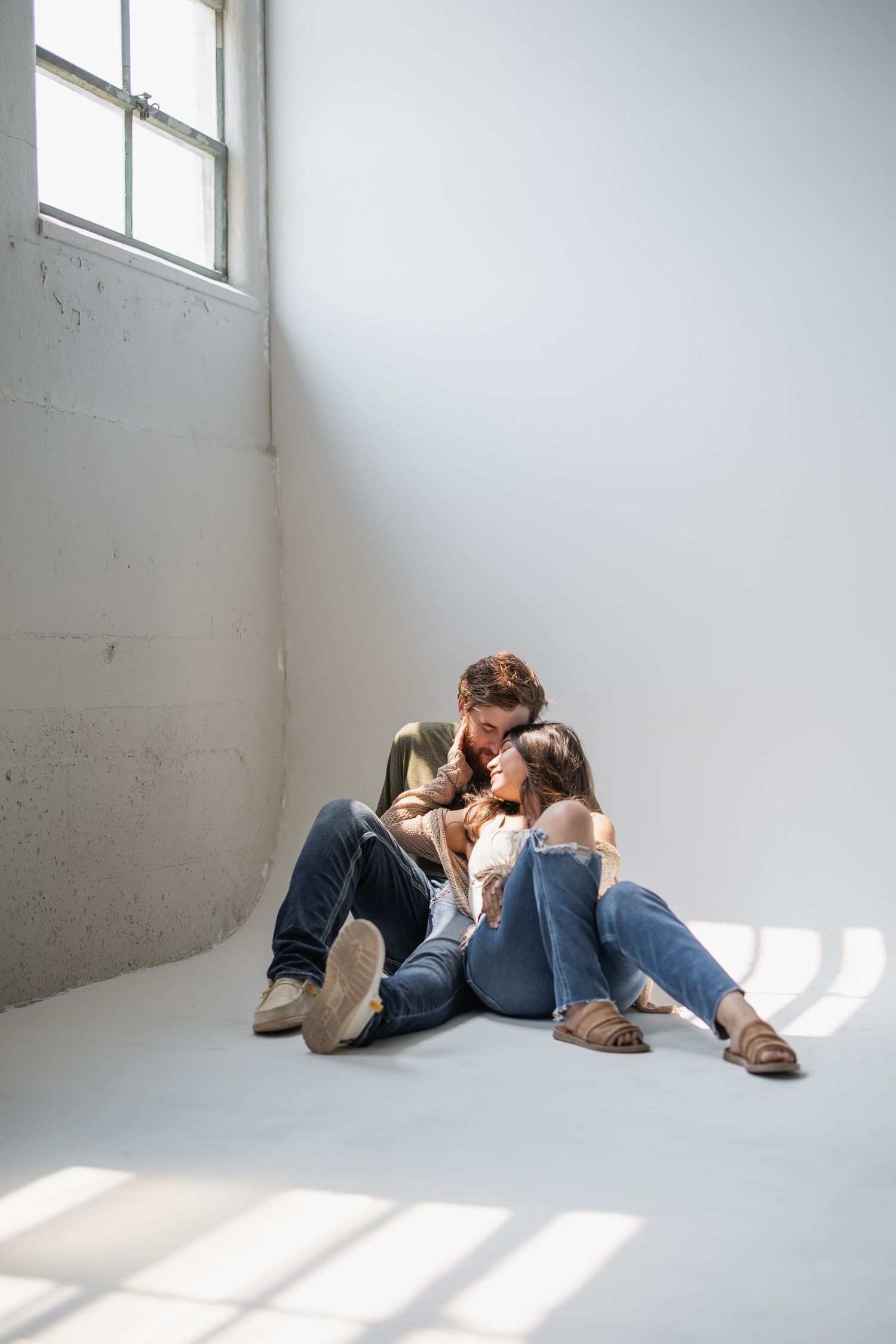 A couple lying on a white floor in a minimalist room with sunlight streaming through windows.