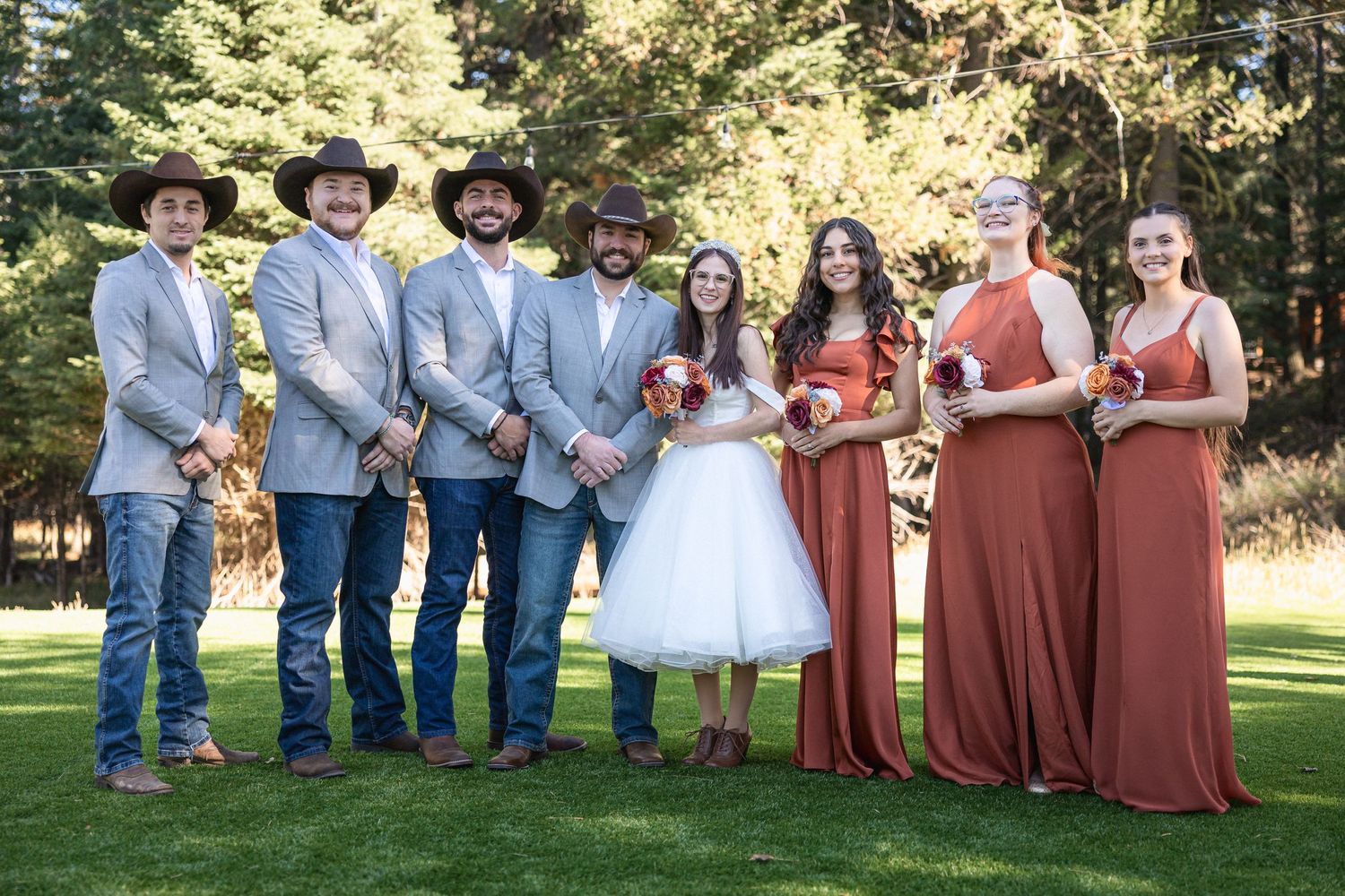 Wedding party poses outdoors with groomsmen in light blue suits and cowboy hats alongside bridesmaids in rust-colored dresses.