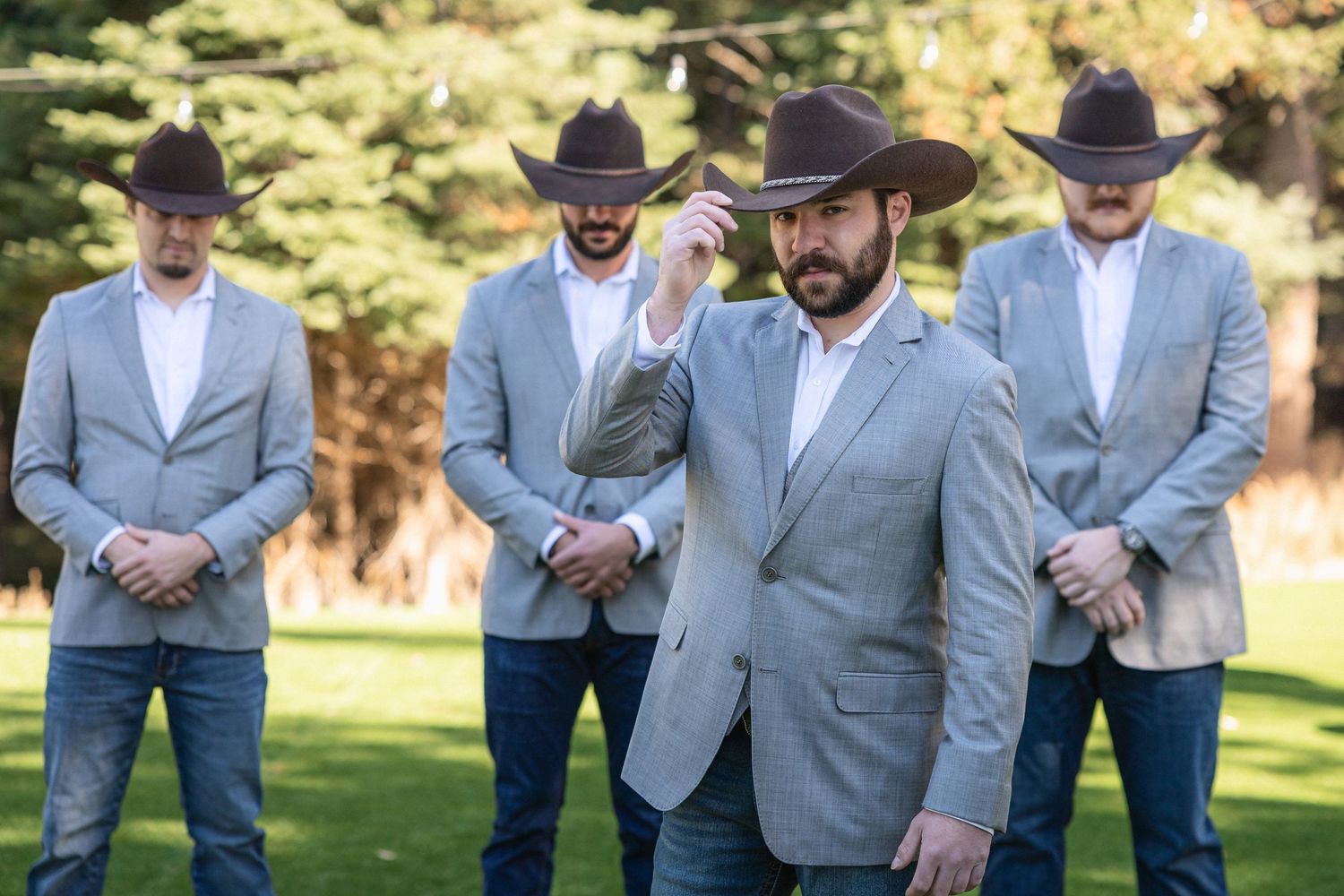 Groomsmen in matching gray suits and cowboy hats standing on lawn at outdoor wedding.