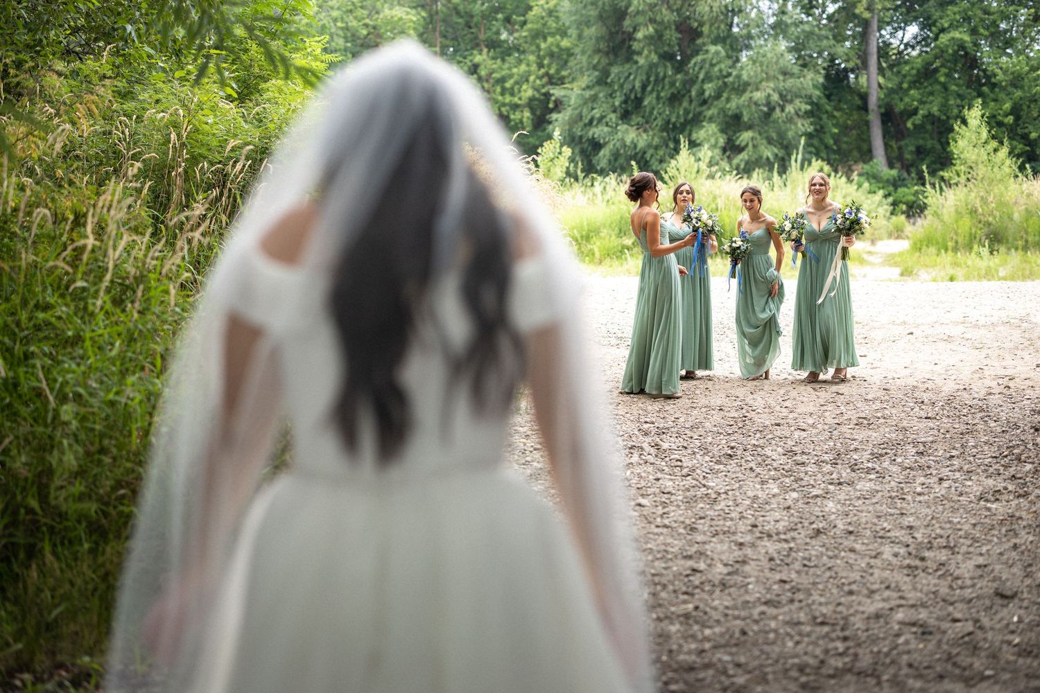 Wedding party members in sage green dresses and navy suits stand together on a wooded path.