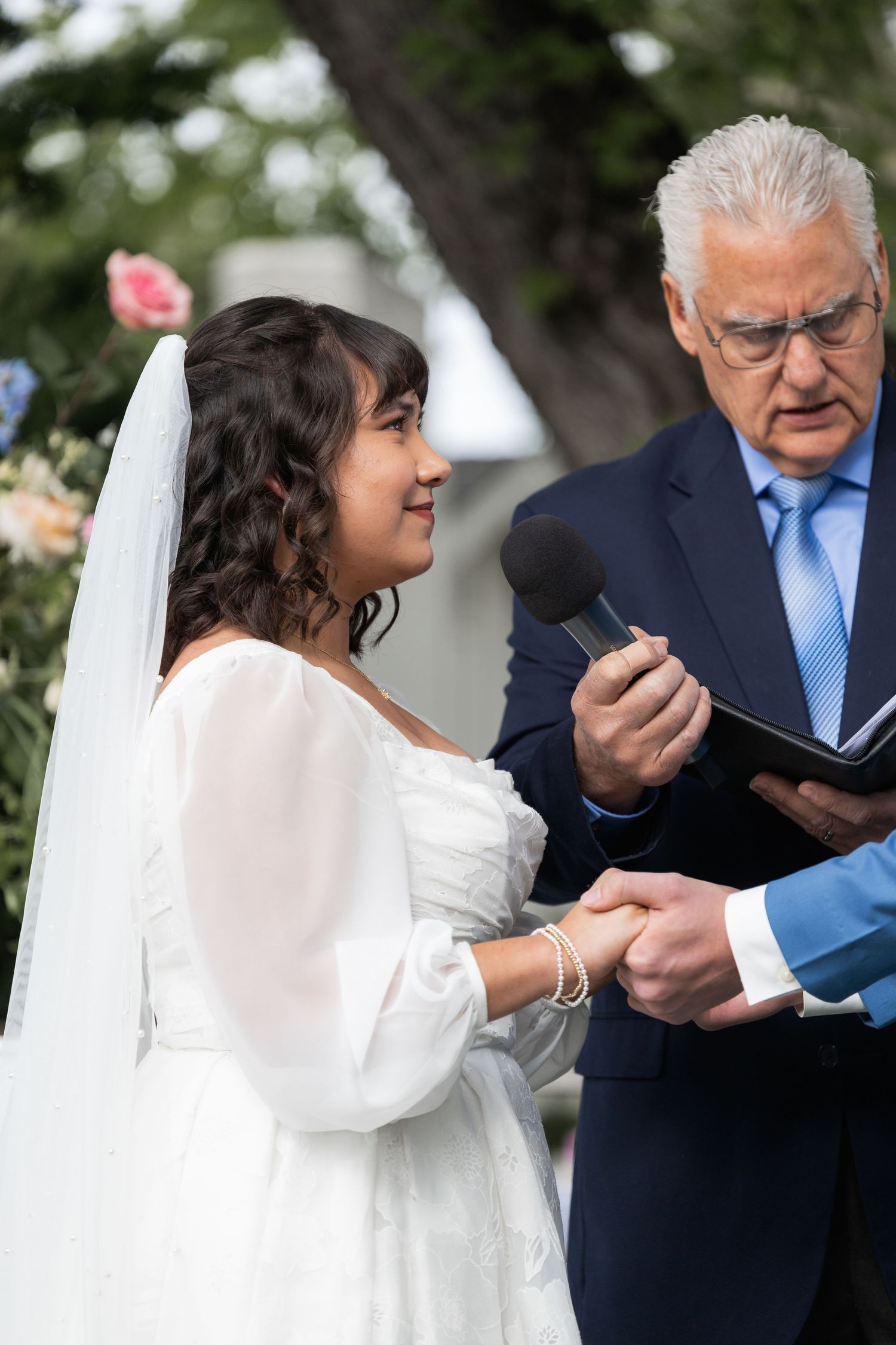 Wedding officiant reads from book during outdoor ceremony under flowering tree.