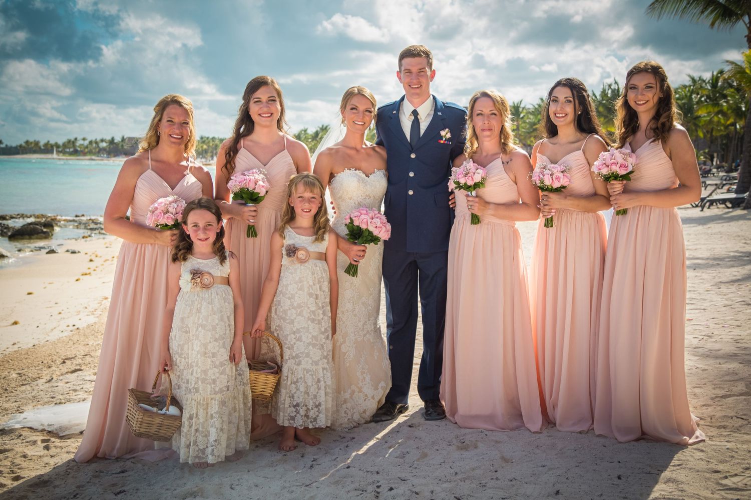 Wedding party poses on a tropical beach with bridesmaids in flowing pink dresses and groom in navy suit.