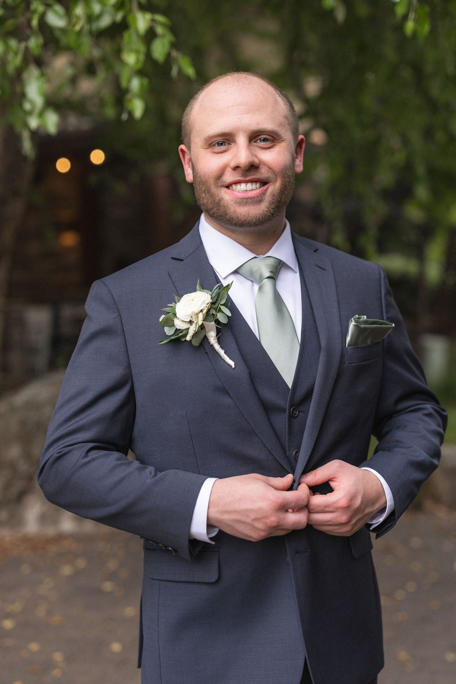 Wedding day portrait of groomsman in charcoal suit with boutonniere against leafy background.
