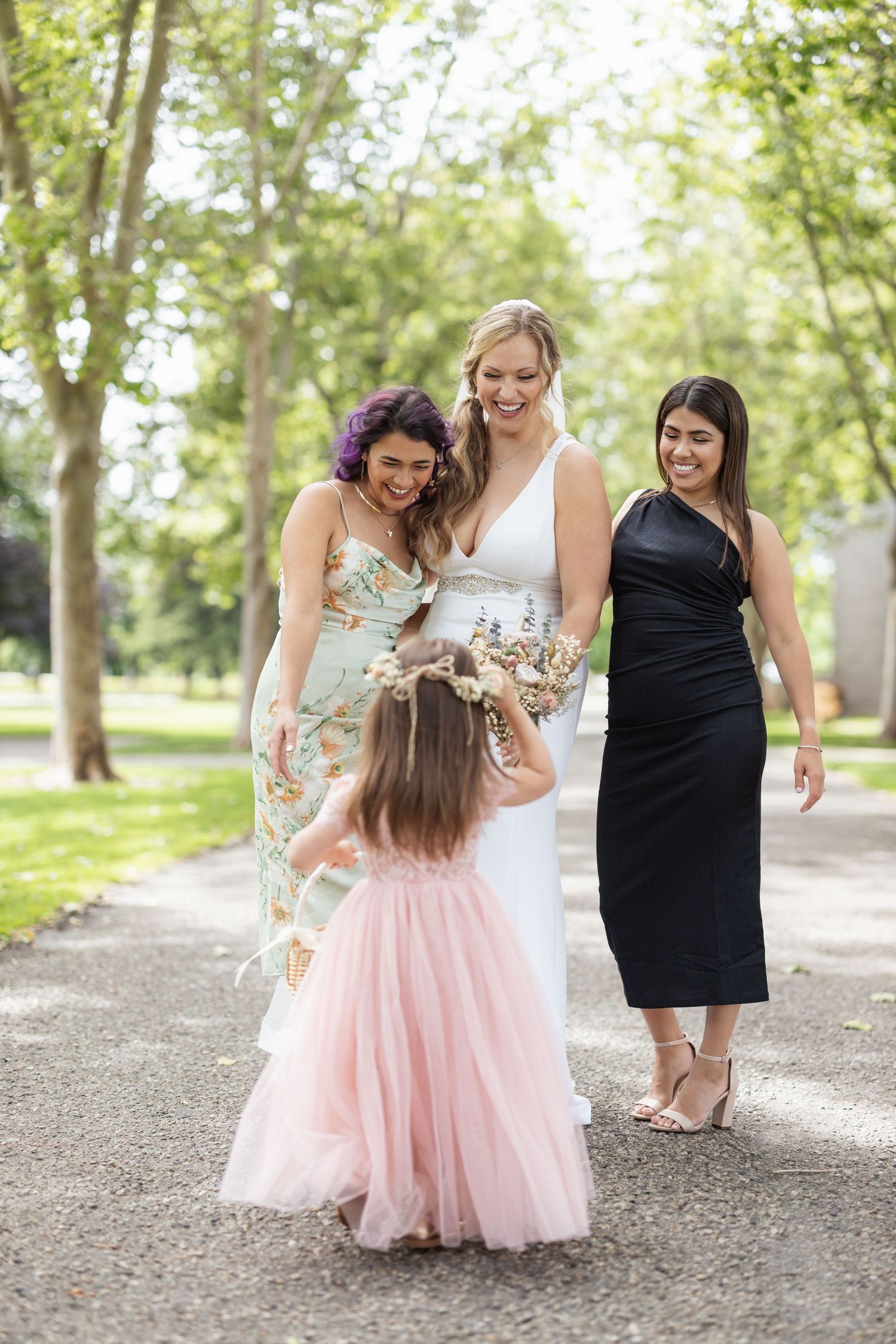 Bride in white dress interacts with young flower girl while two women in formal attire watch.