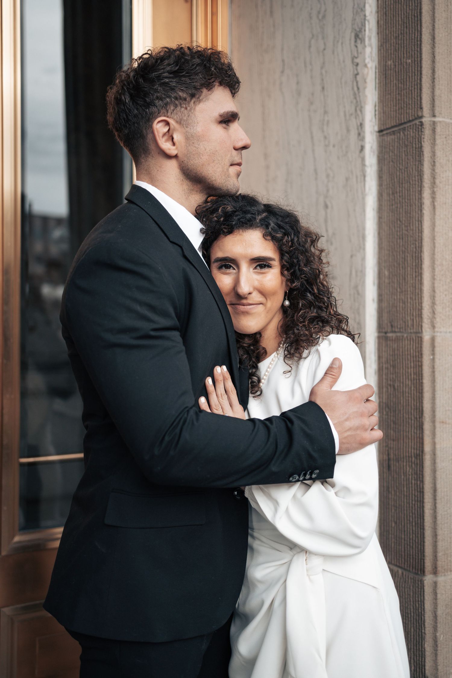 A couple in formal attire embrace lovingly against a stone building facade.