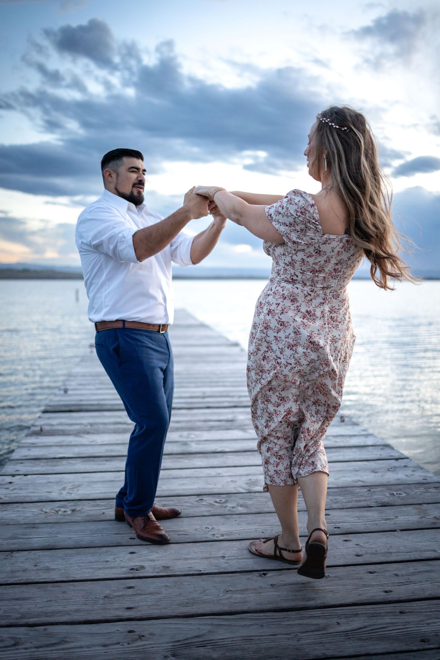 A couple dances joyfully on a wooden dock during sunset with dramatic clouds overhead.