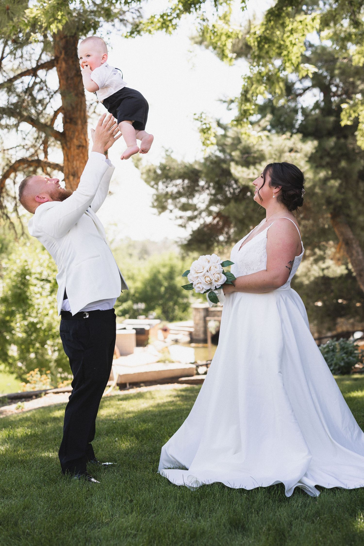Playful moment at a wedding as a person lifts a small child while the bride looks on with her bouquet.