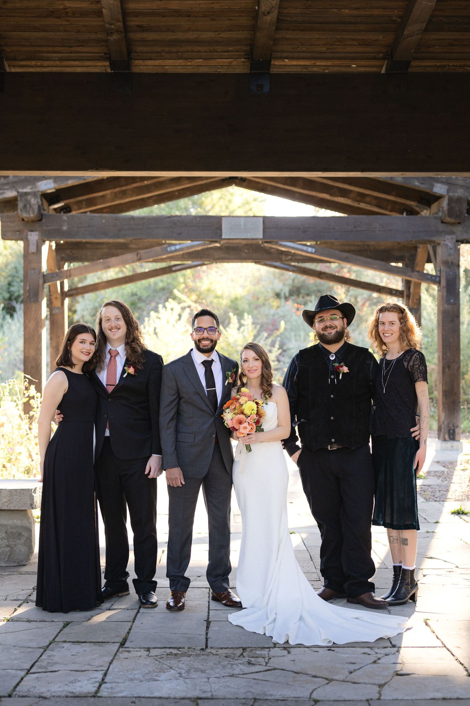 Wedding group photo showing couple and attendants in black attire under wooden pergola.