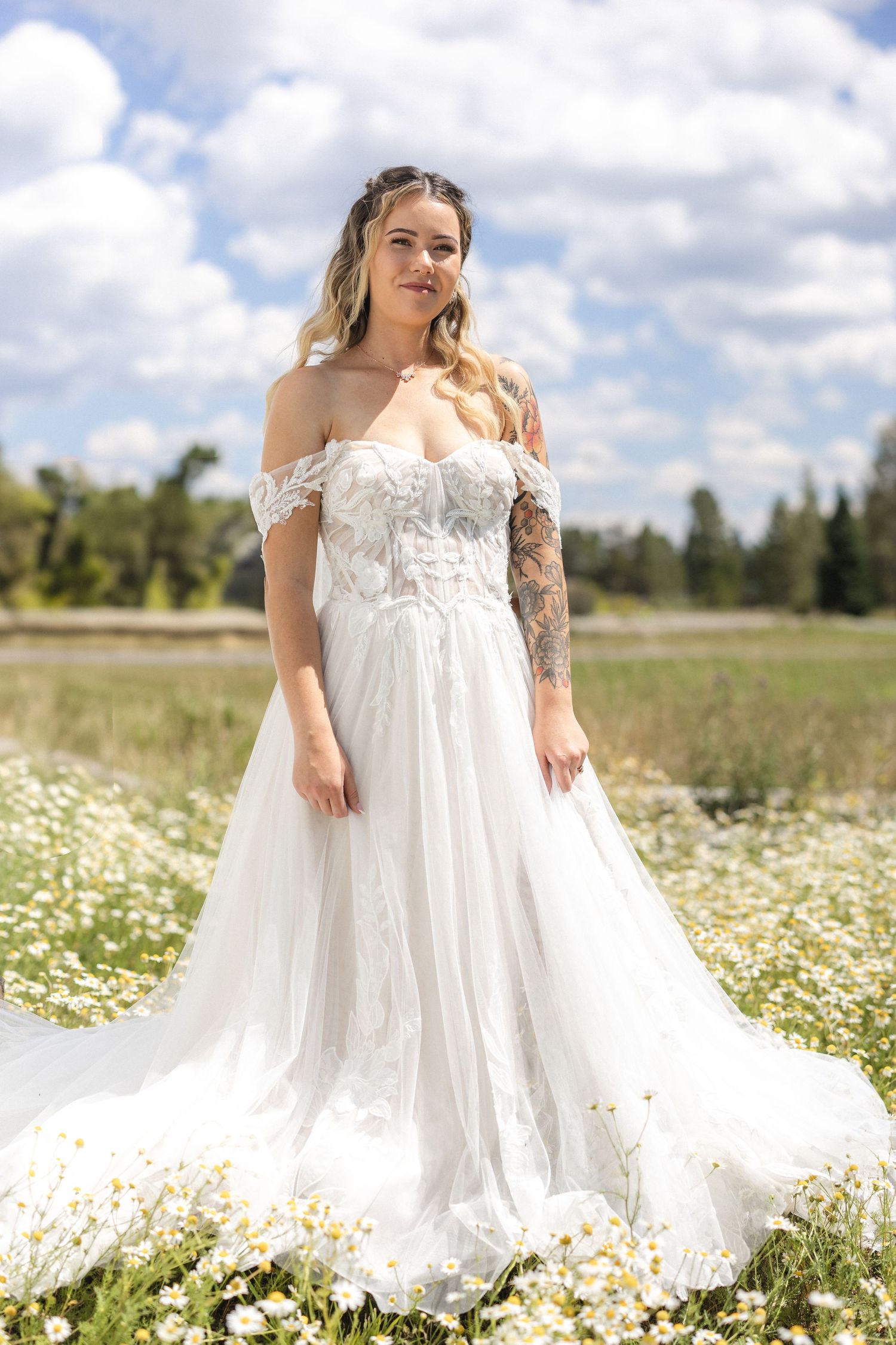 A bride in an off-shoulder lace wedding gown poses in a field of blooming daisies.
