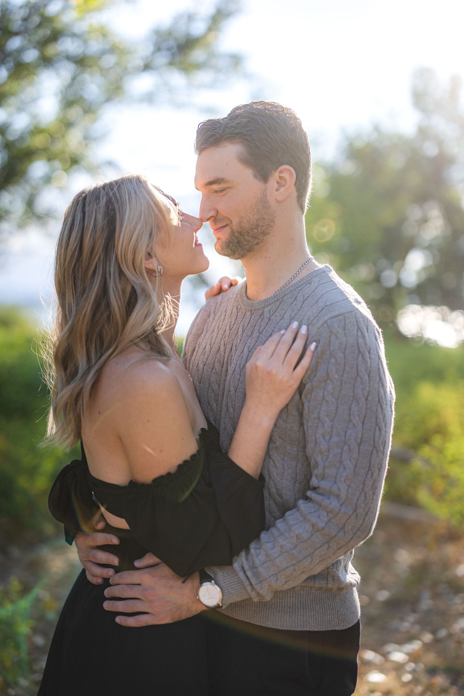 Romantic couple embracing outdoors in sunlit natural setting with woman in black dress and man in grey sweater.