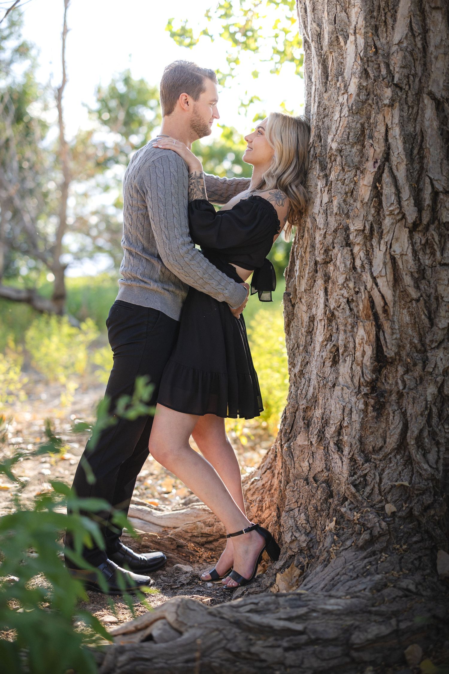 Intimate moment between couple in black dress and gray sweater against large tree trunk.