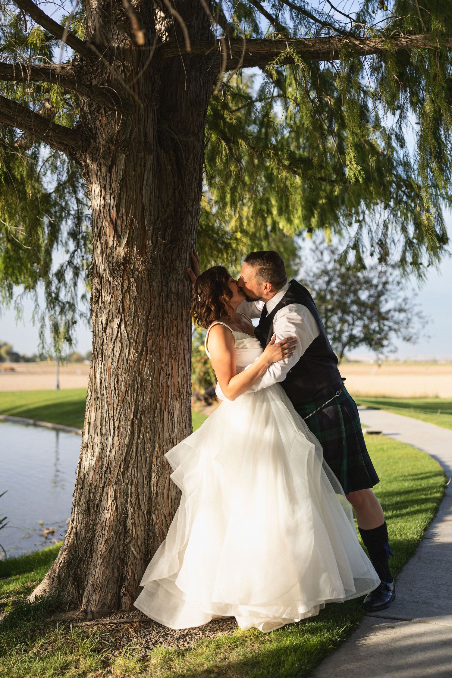A couple embraces under a large tree beside a pond during their wedding photoshoot.