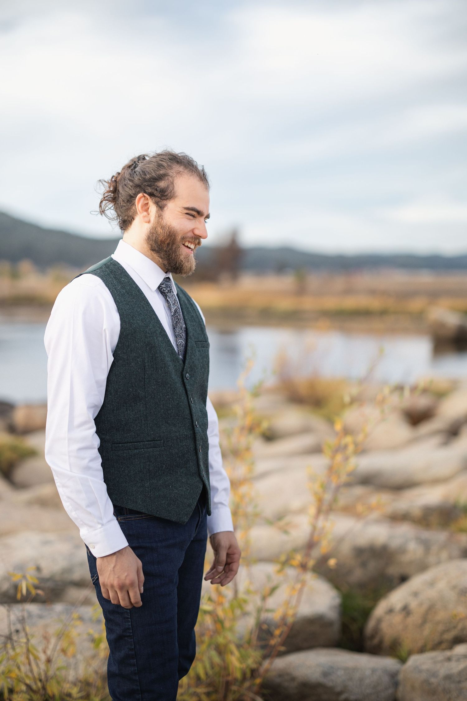 Person in formal vest and white shirt standing on rocky terrain near water with mountains in background.