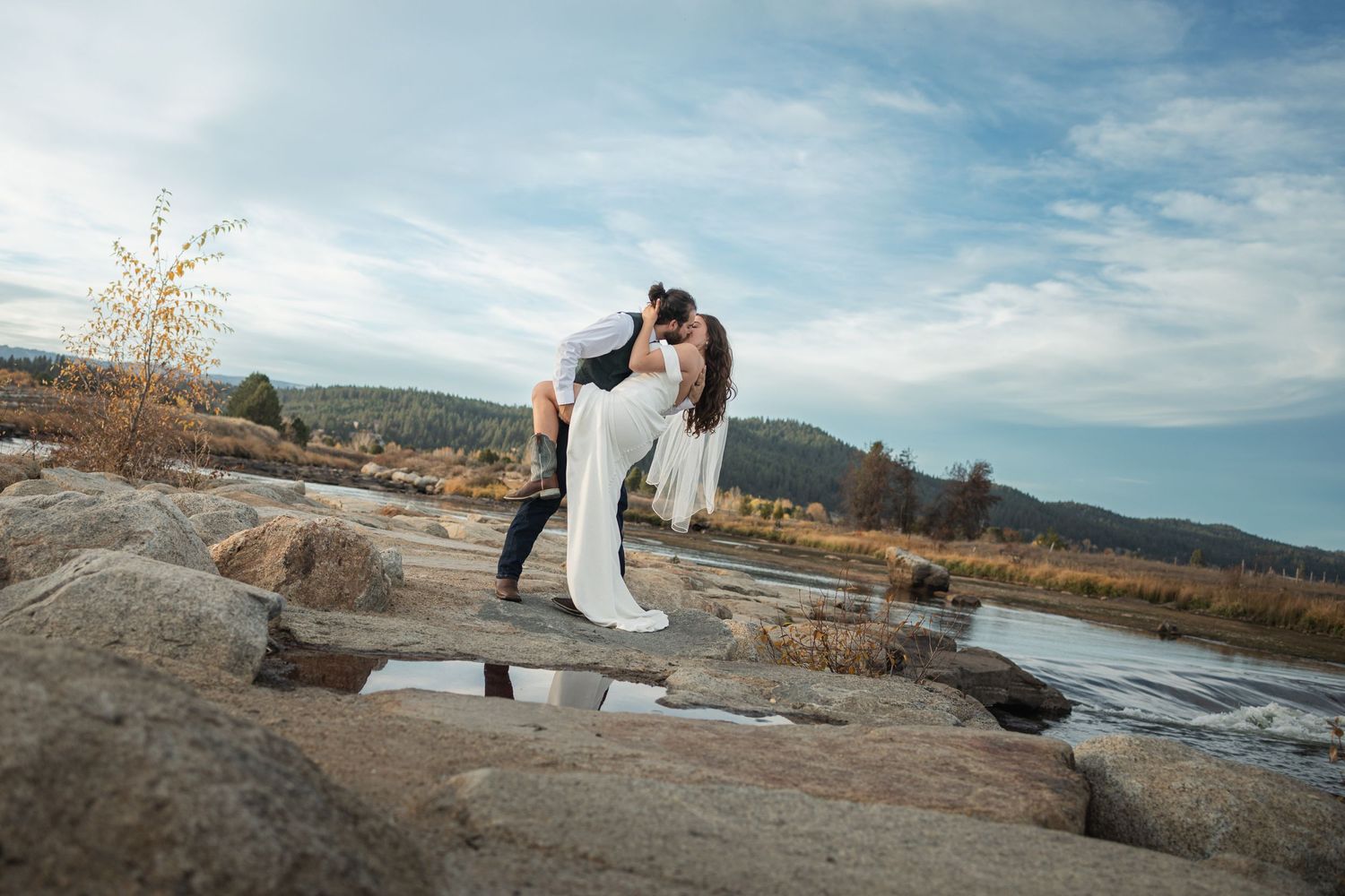 Wedding couple shares intimate moment on rocks by rushing river with mountain landscape.