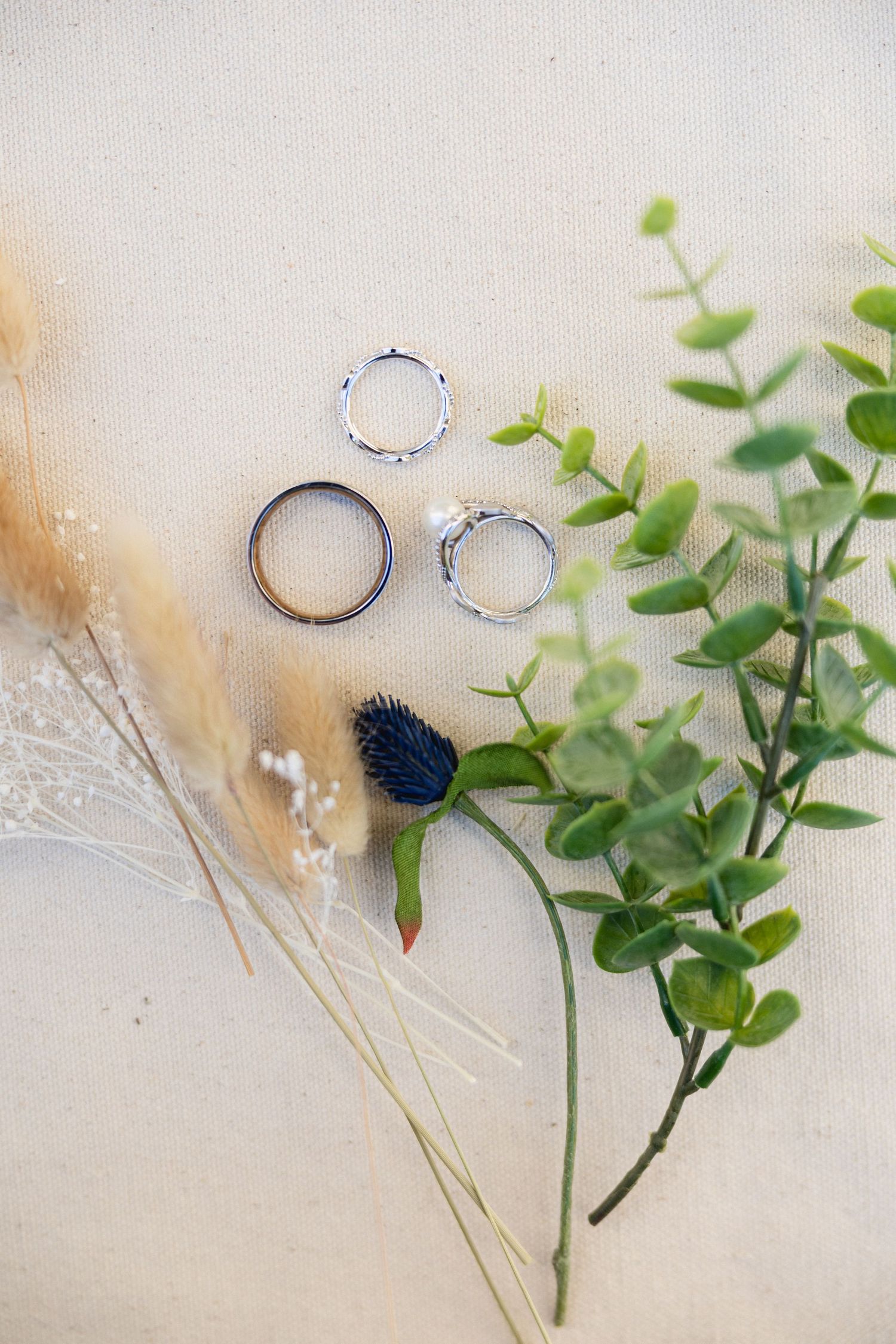 Three wedding bands arranged with greenery and dried flowers on white surface.