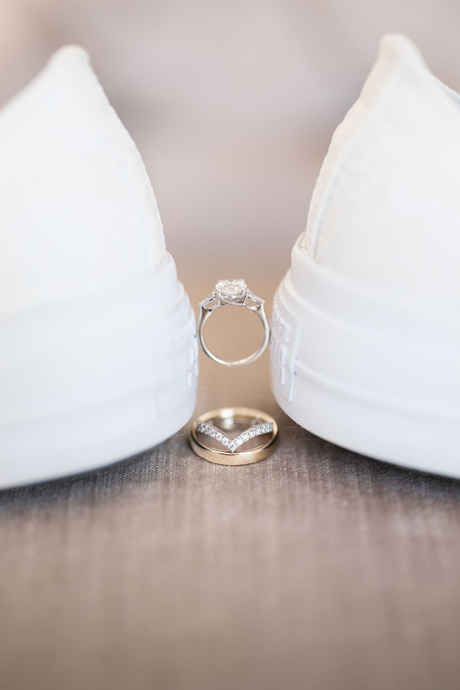 Wedding rings artistically placed between white bridal shoes on wooden surface.