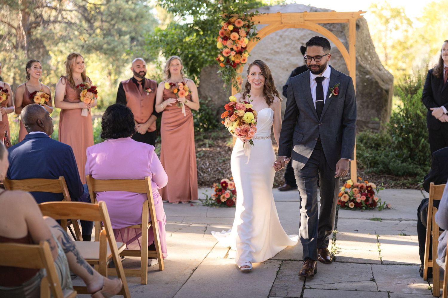 Wedding guests seated in wooden chairs watching outdoor ceremony under covered venue.
