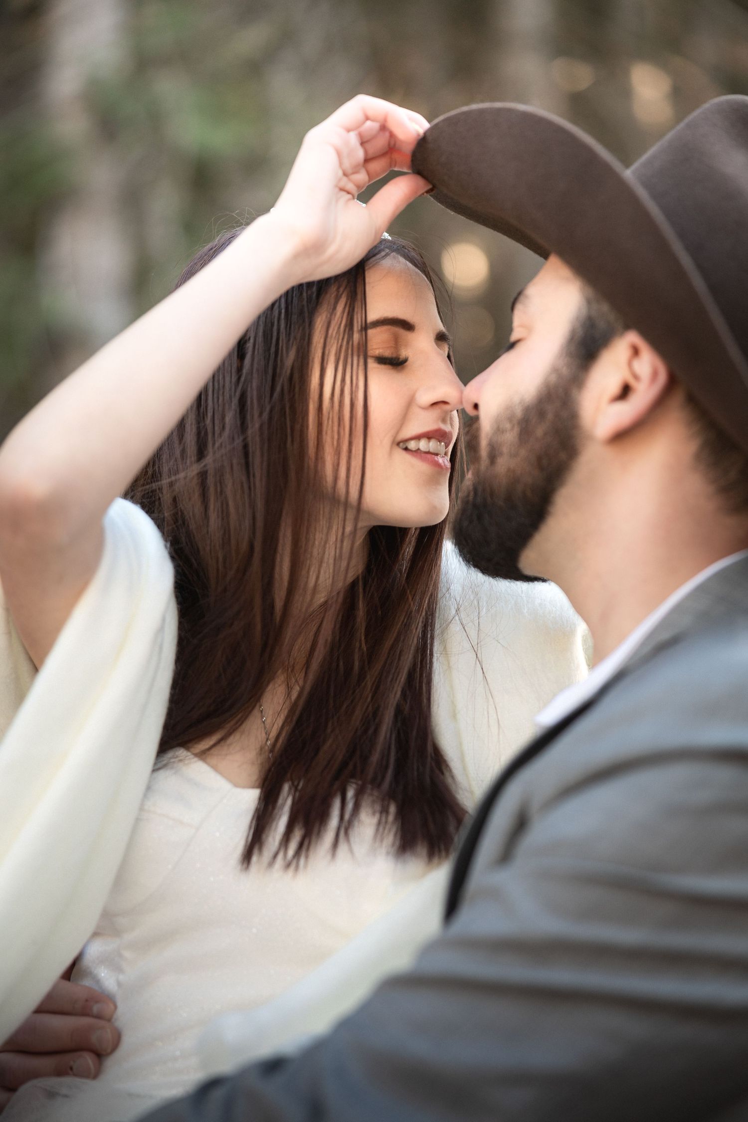 A tender moment between a couple in wedding attire with a cowboy hat against a forest backdrop.