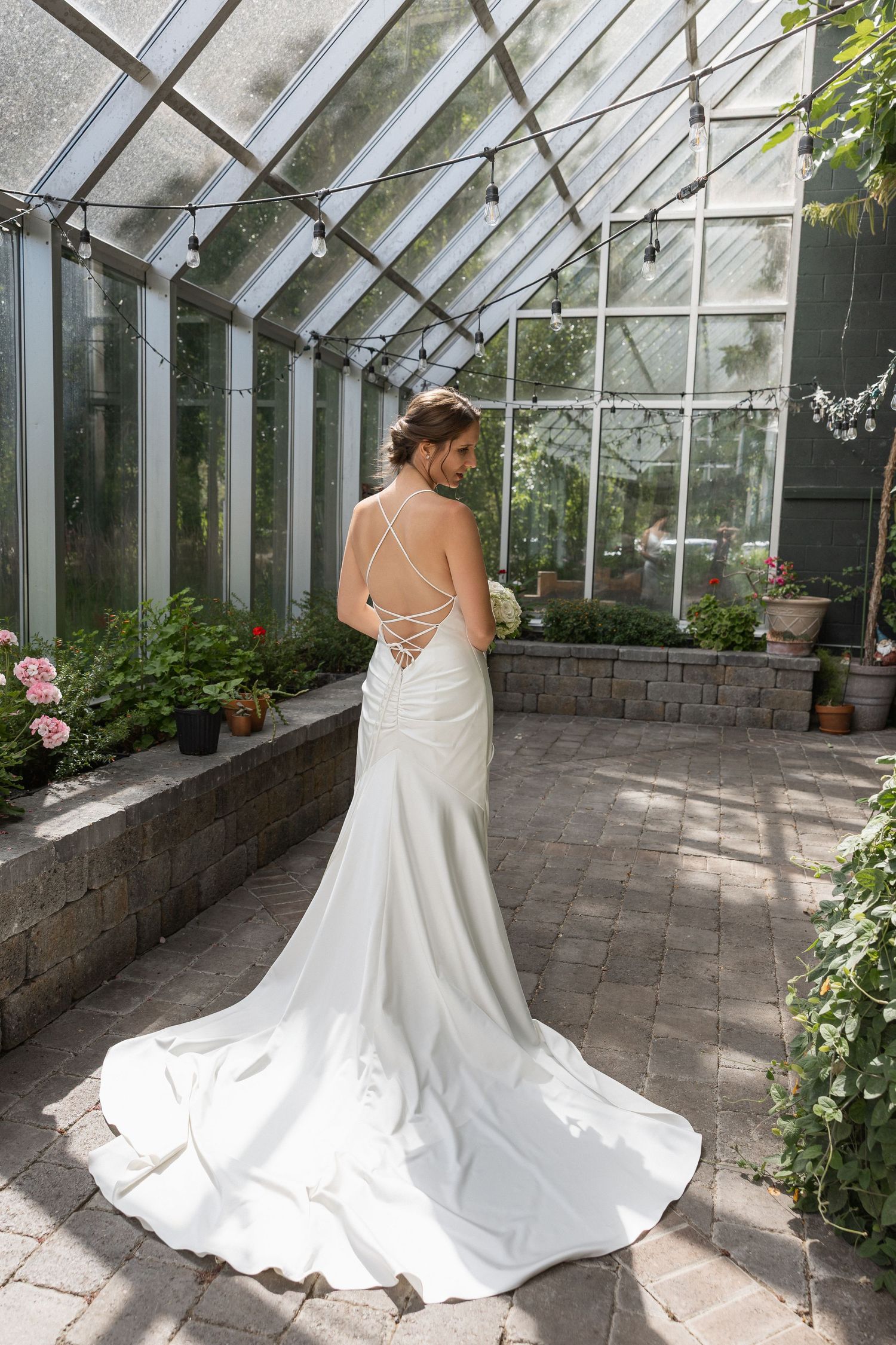 Elegant white wedding dress with flowing train photographed in a sunlit greenhouse conservatory.