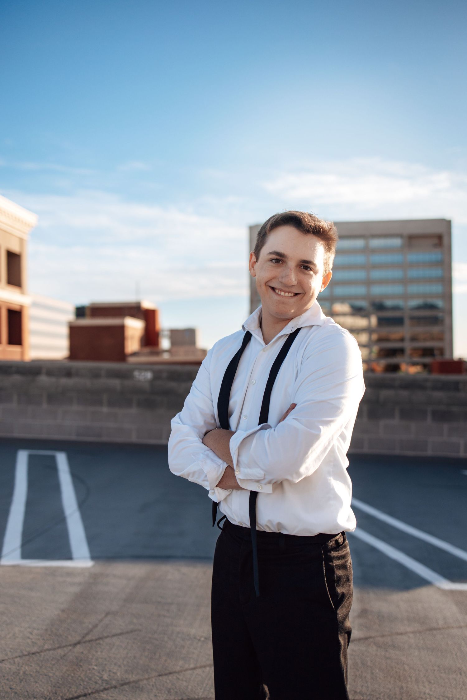 Person in white dress shirt and suspenders standing on rooftop parking area with urban buildings in background.