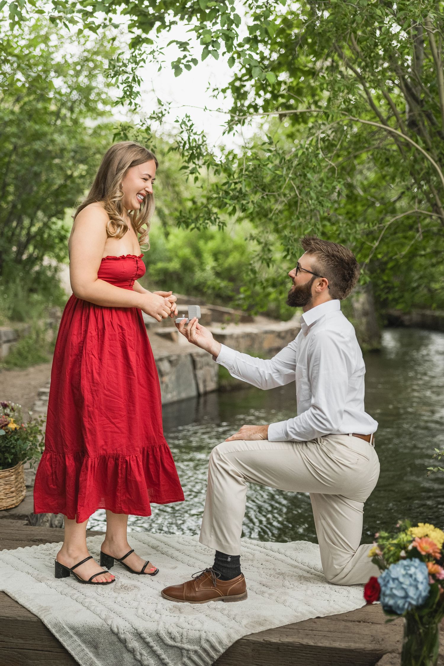 Man kneeling on one knee proposing to woman in red dress outdoors near a creek with lush green trees.