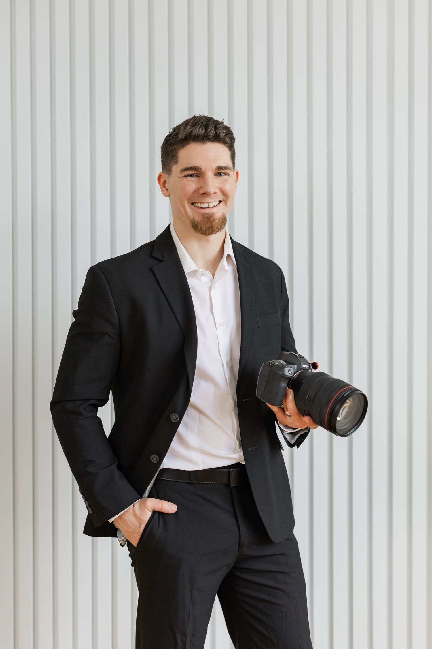 Professional wearing a black suit and white shirt poses casually against a white striped wall background.