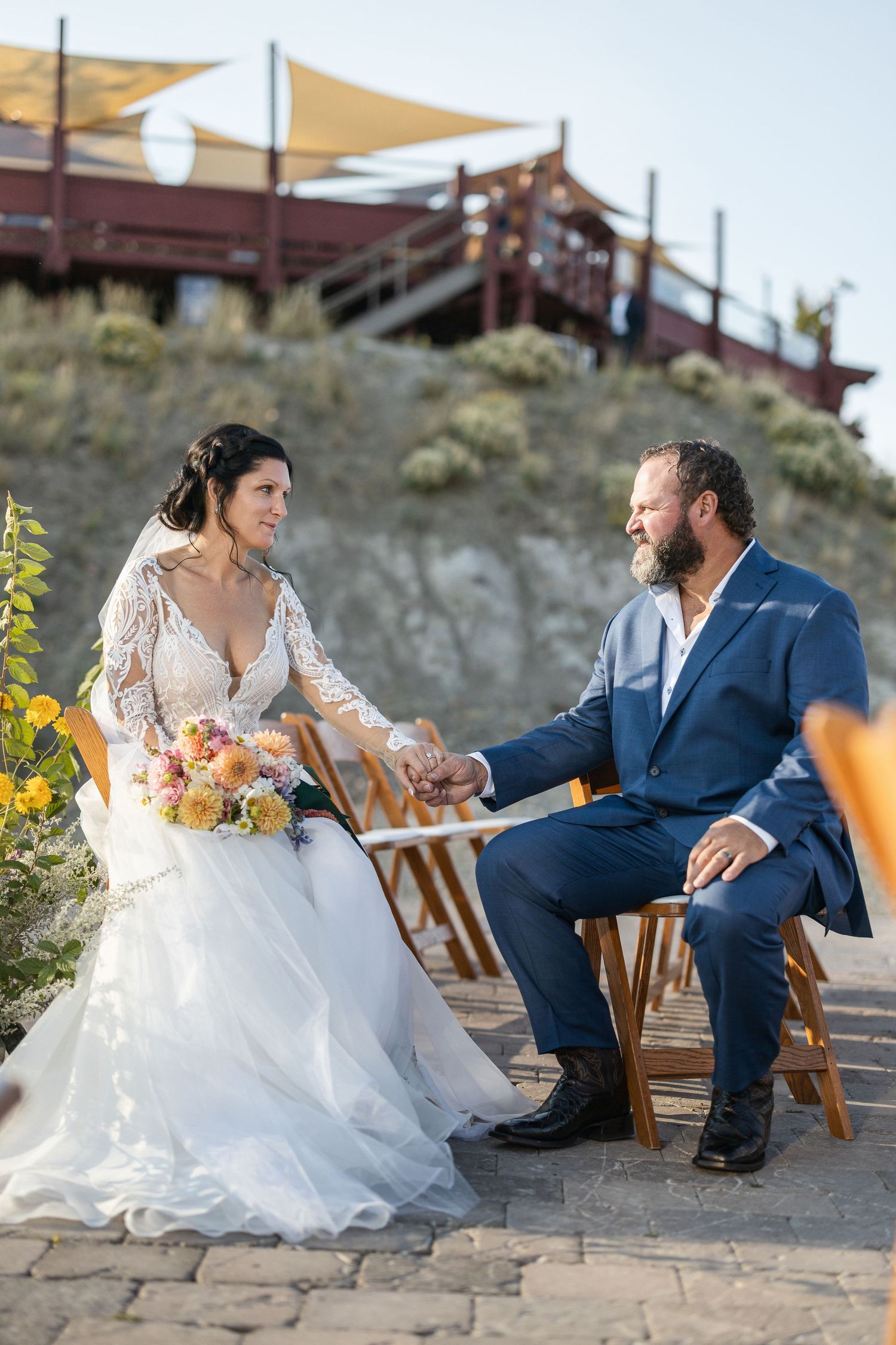 Couple in wedding attire sitting and laughing together on wooden chairs at an outdoor venue.