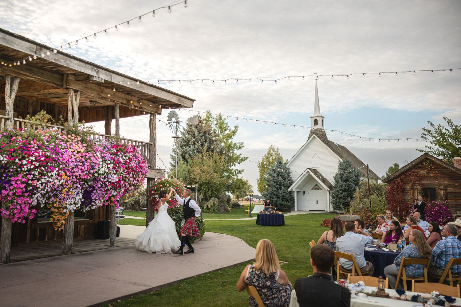 Outdoor wedding reception with string lights, white chapel, and colorful flowers in rustic venue.