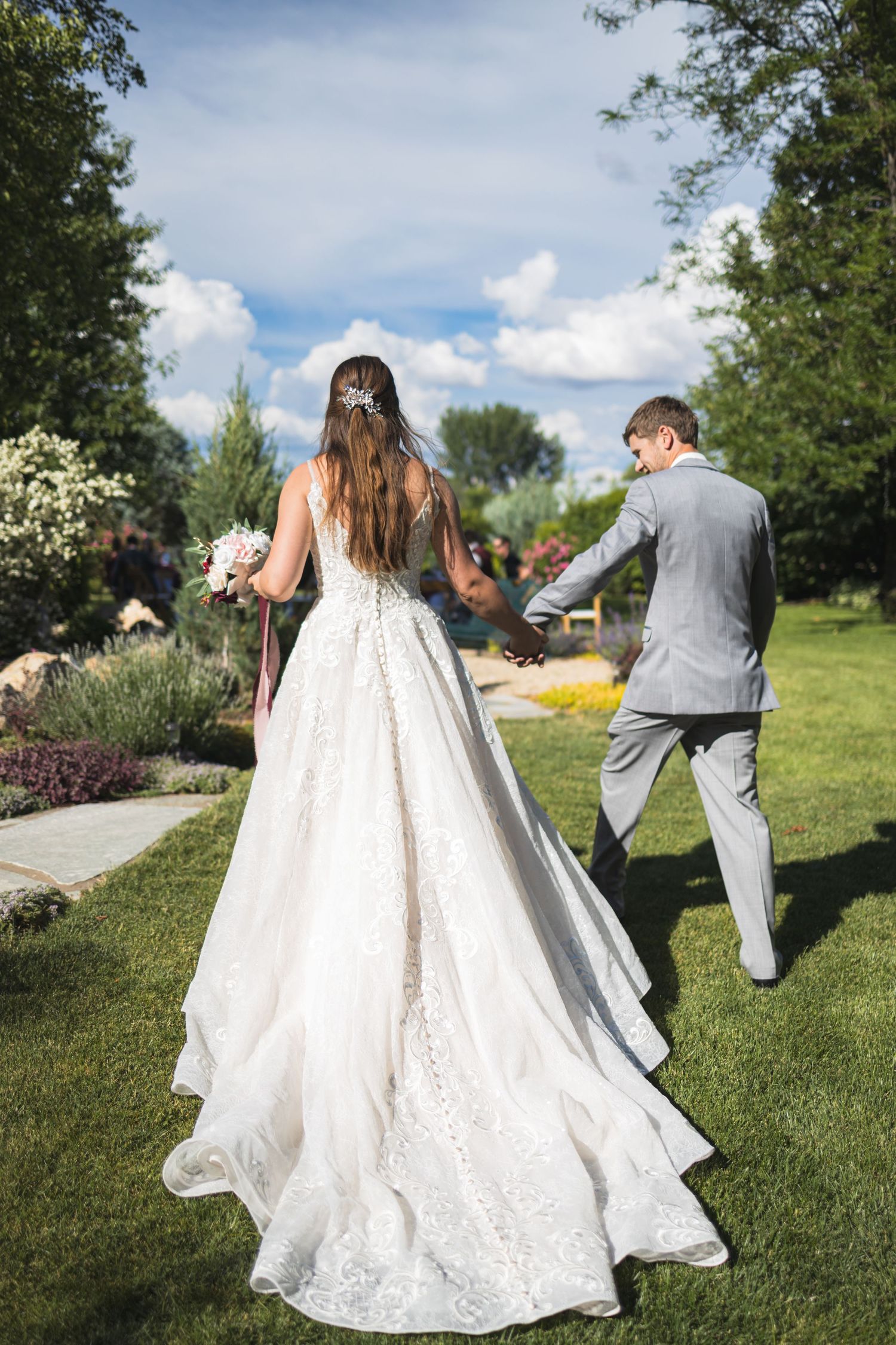 A wedding couple walking through a garden path, showcasing the beautiful train of the bride's dress.