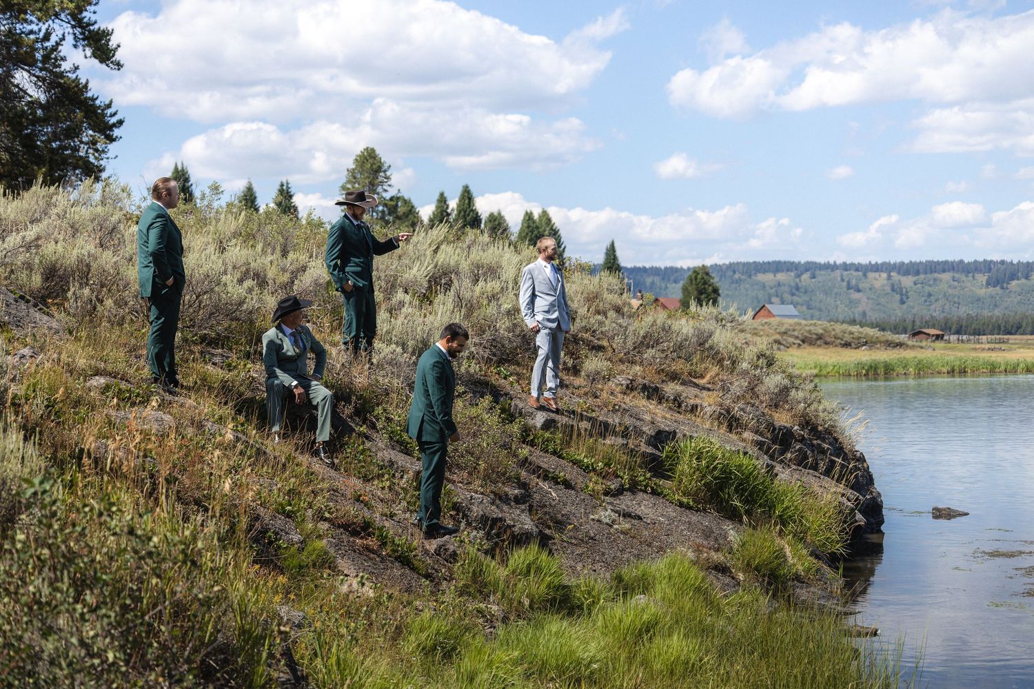 Group of people standing on grassy hillside overlooking serene lake on sunny day.
