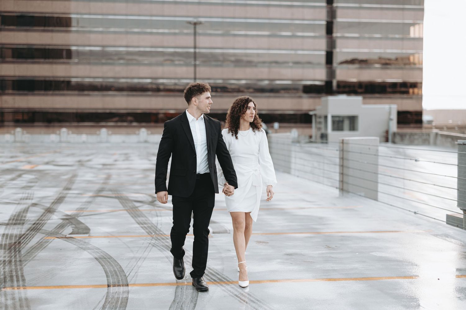 A couple in formal attire walks together on an urban rooftop parking structure.