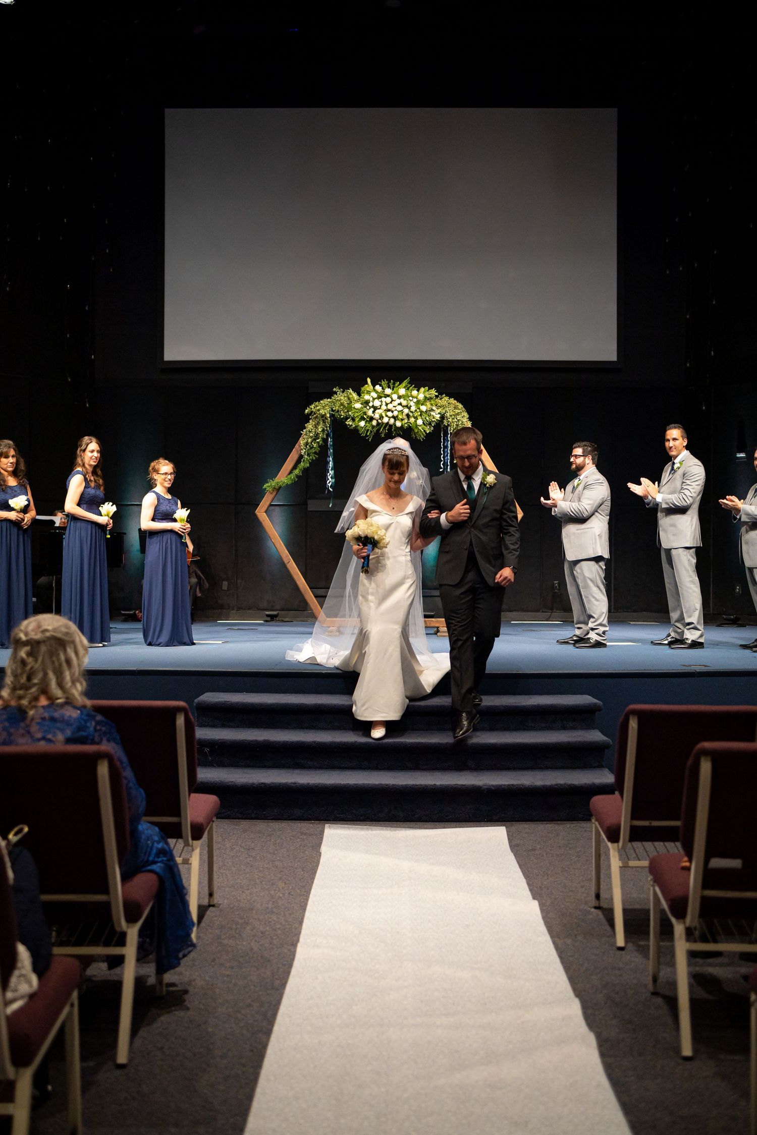 Wedding recessional down white aisle with geometric arch and navy bridesmaids in background.