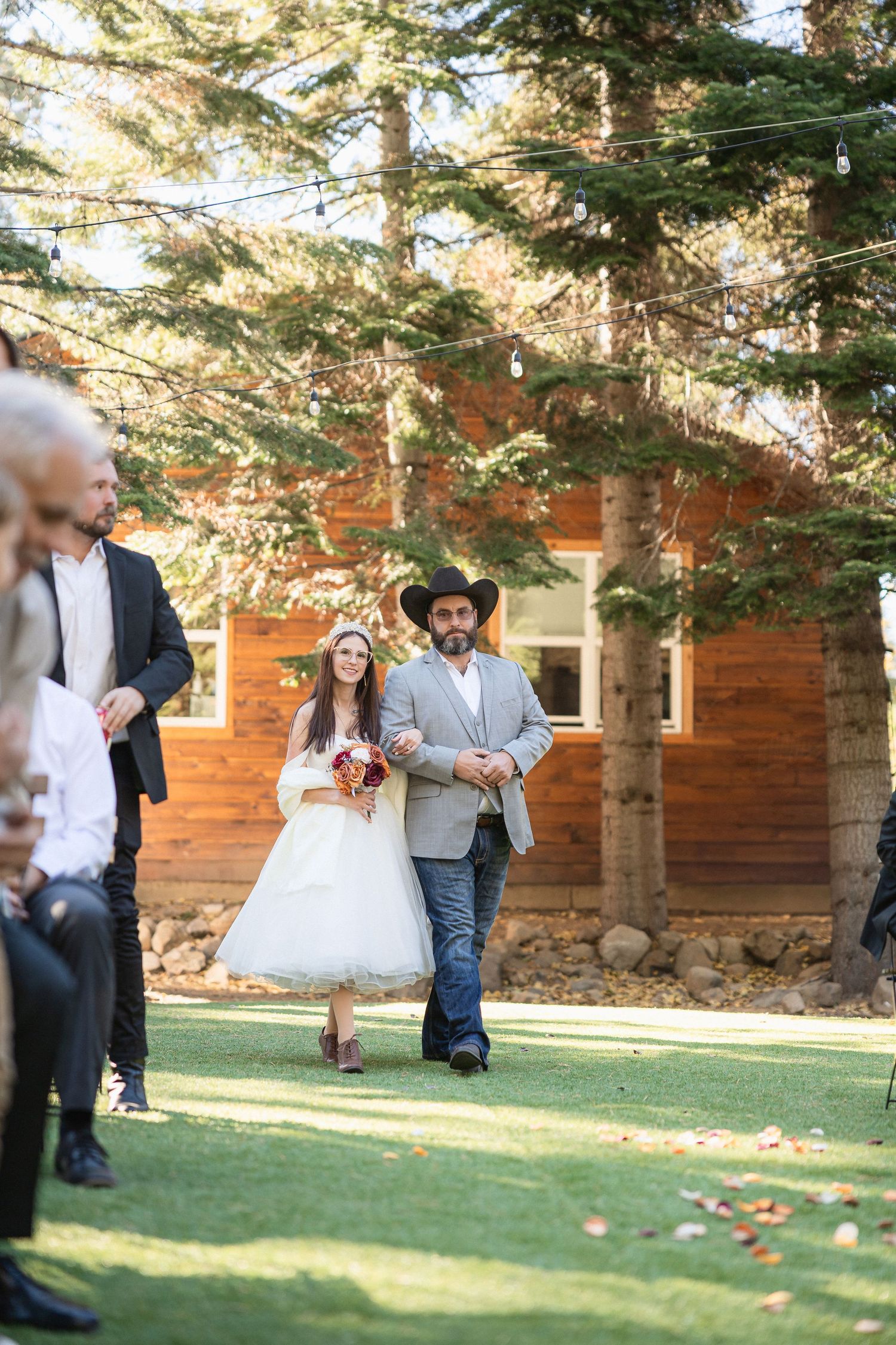 Wedding party walking on a grassy lawn near a rustic wooden cabin with evergreen trees in the background.