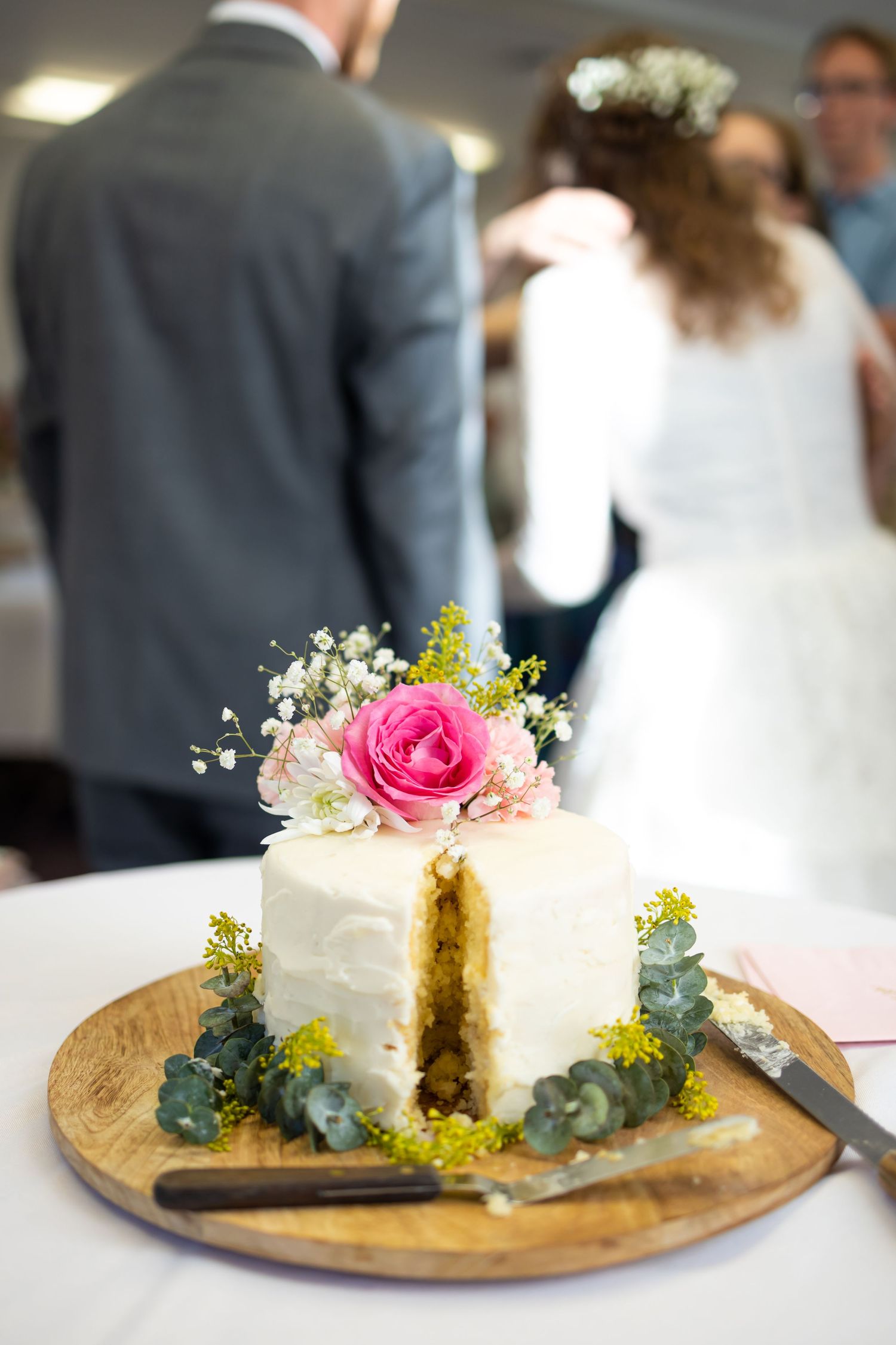 A wedding cake with fresh flowers and eucalyptus on display with the couple in the background.