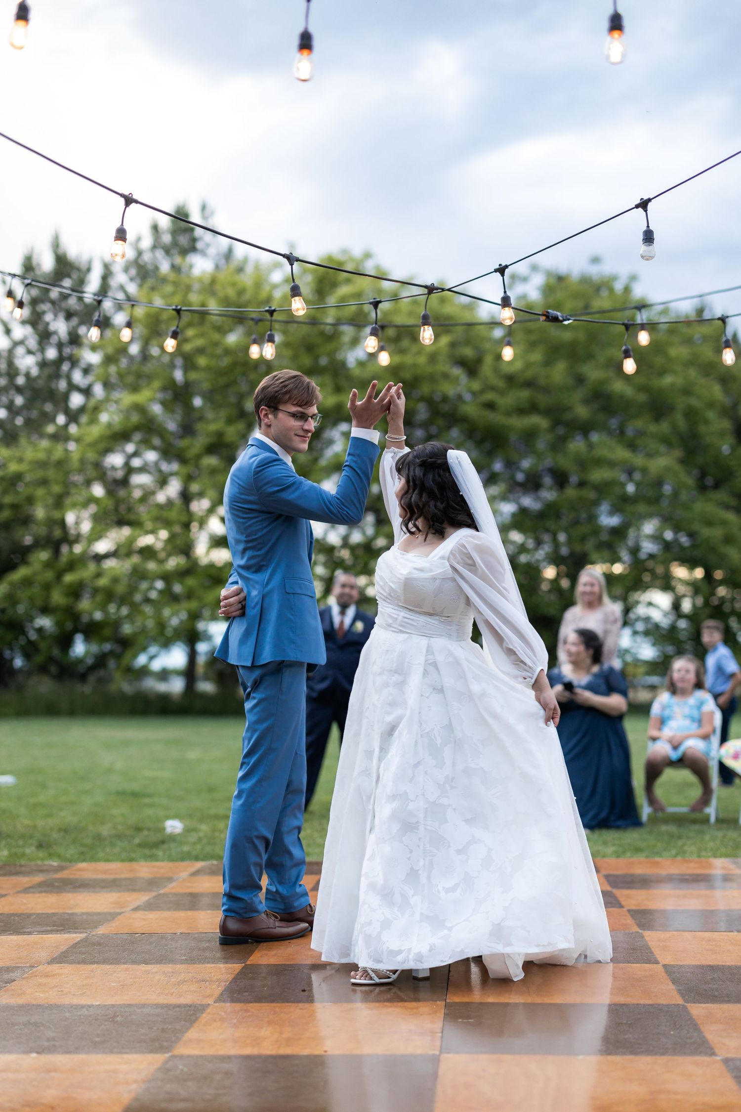 Couple dances on checkered dance floor under string lights at outdoor wedding reception.