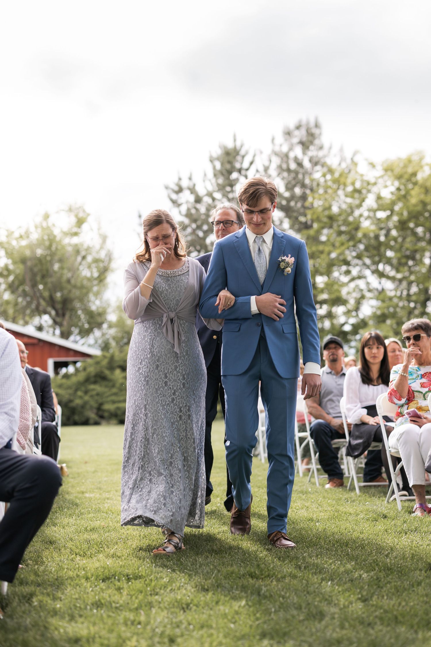 An outdoor wedding processional on a grassy lawn with guests seated in white chairs.