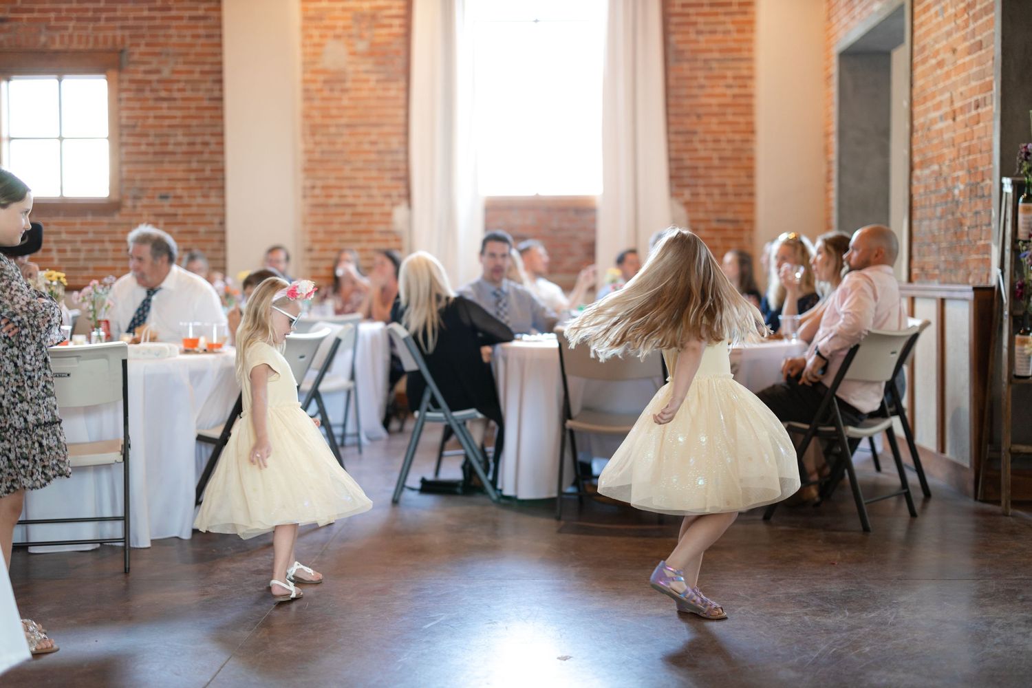 Young flower girls in white dresses twirl on dance floor during wedding reception.