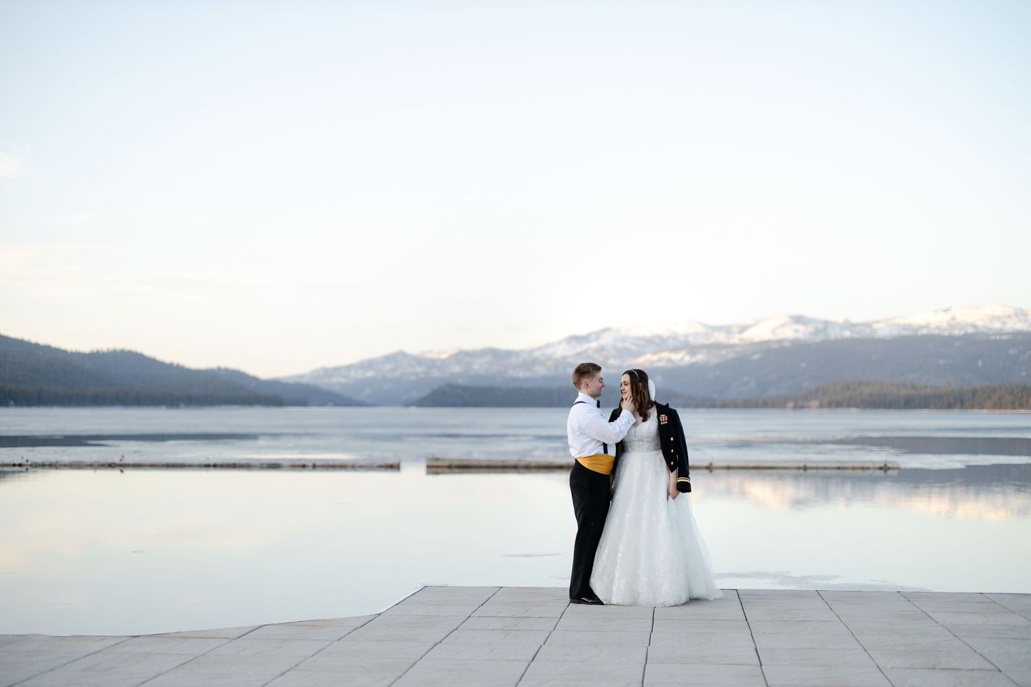 A couple in formal wedding attire poses for romantic photos against a serene mountain lake backdrop at sunset.