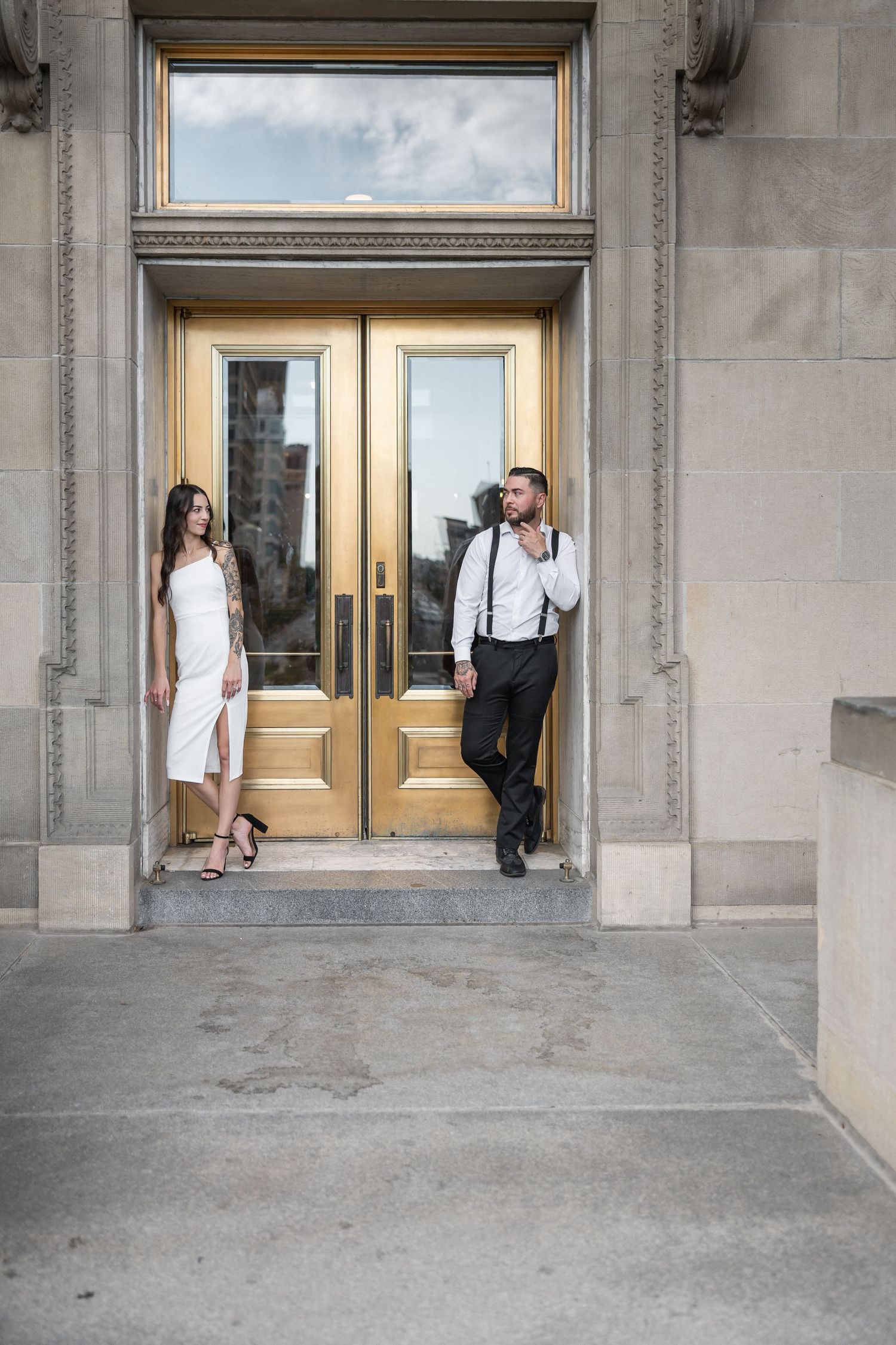 Couple in white and black attire pose romantically by ornate golden doors of a classical building.