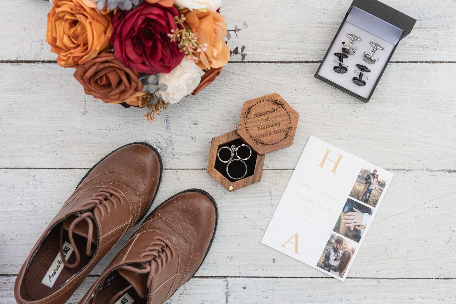 Wedding details flatlay with brown shoes, roses, and ring box on white wooden surface.
