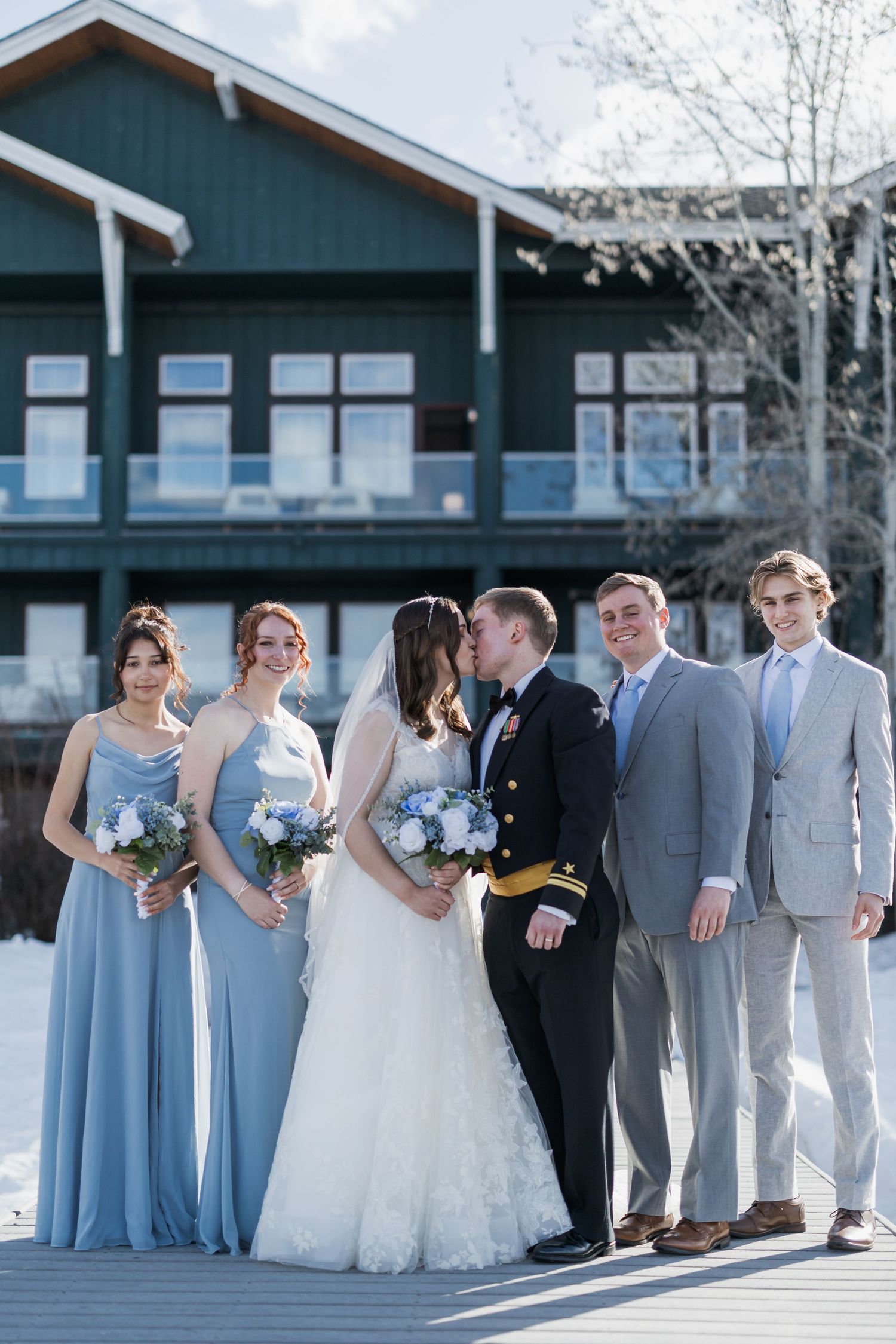 A wedding party poses in front of a blue lodge wearing light blue bridesmaid dresses and gray suits.